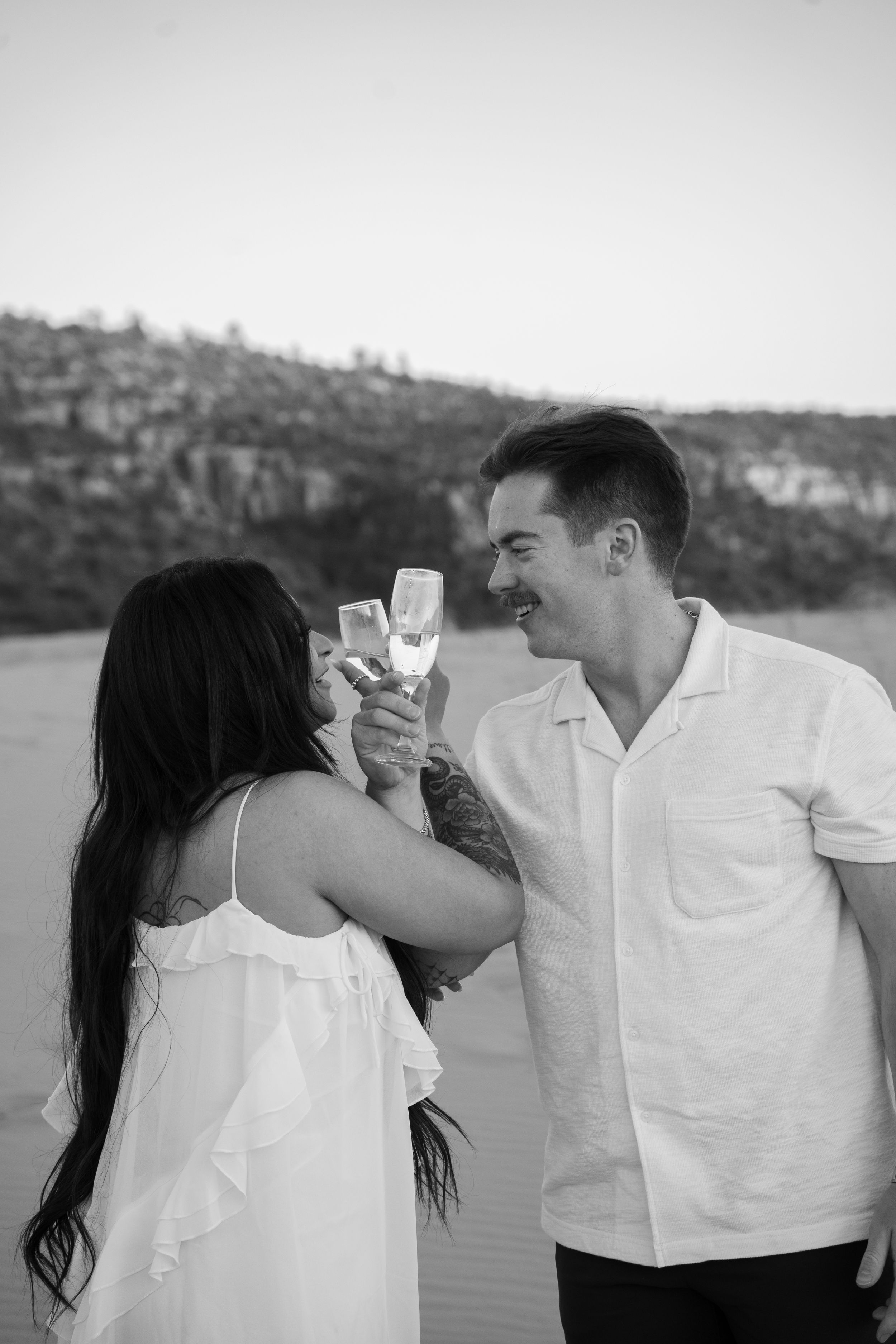 Black and white photo of a couple toasting with champagne glasses on sand dunes, smiling at each other.
