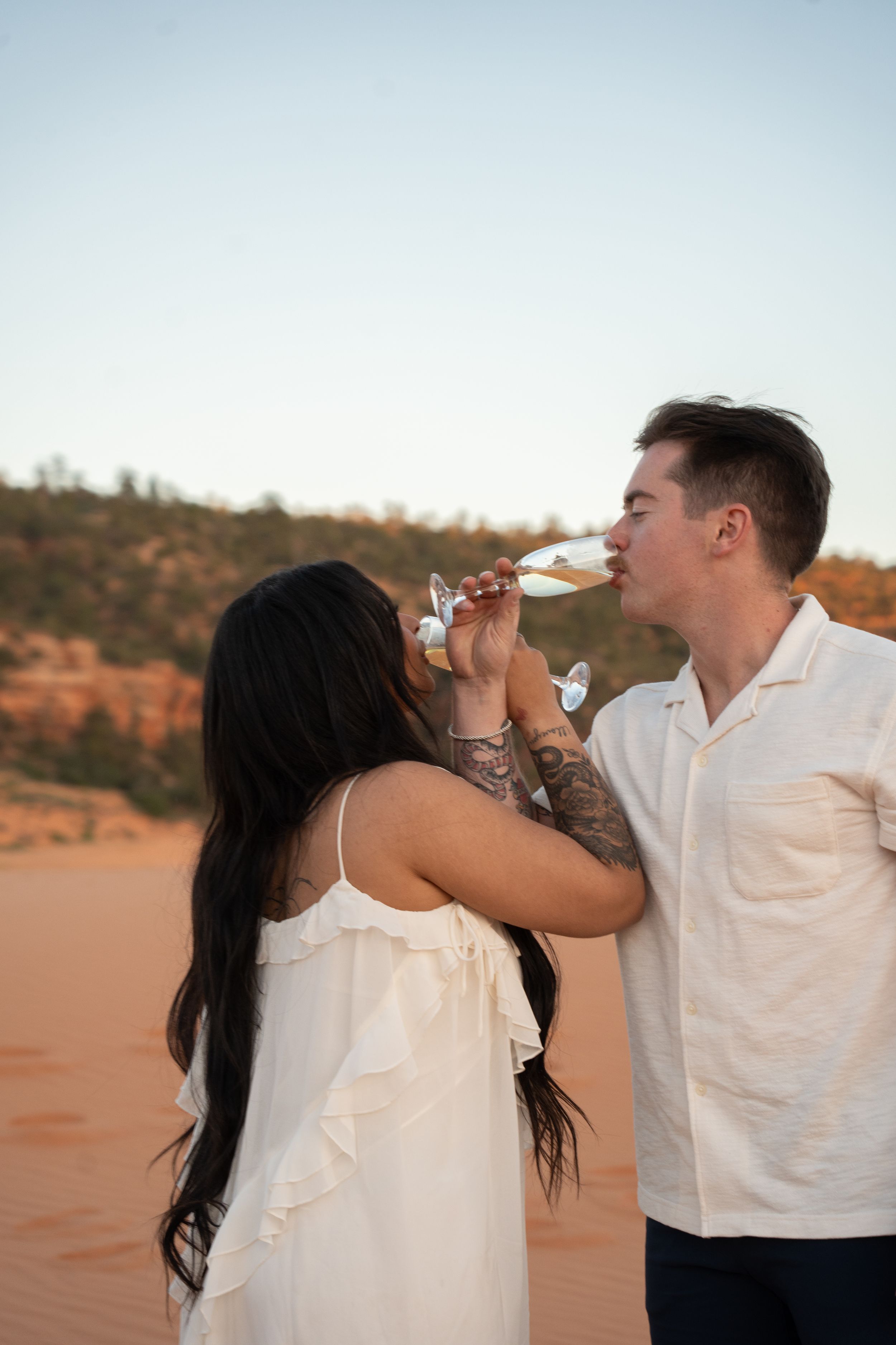 Couple in white outfits drinking champagne with interhooked arms to celebrate their engagement