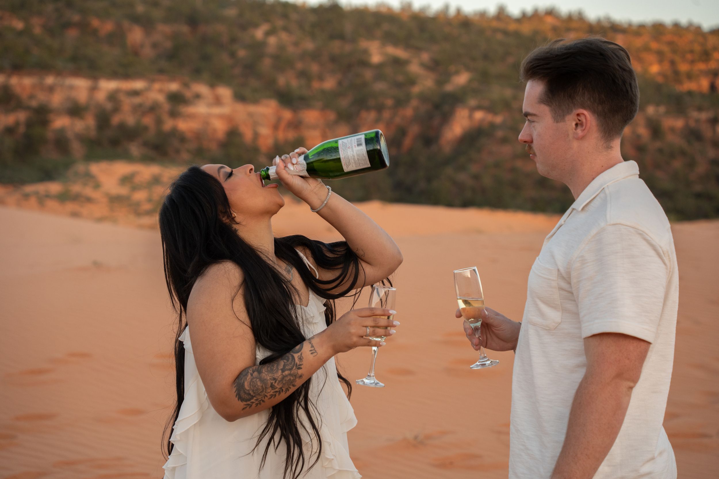 Couple celebrating with champagne in desert landscape, woman drinking from bottle, man holding glass.