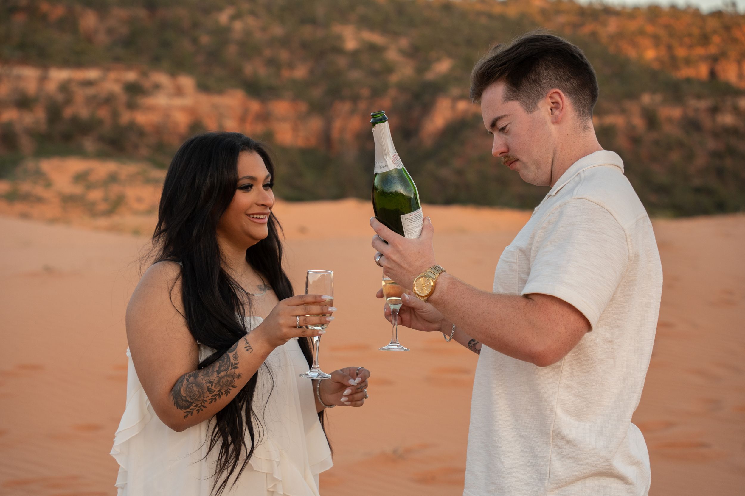 Couple celebrating with champagne on red sand dunes at sunset, woman with tattoos in white dress, man in cream shirt.