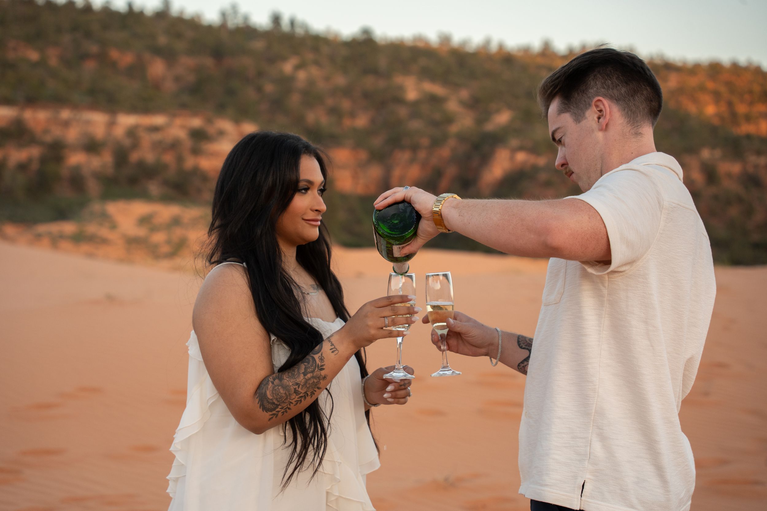 Couple sharing champagne toast in pink sand dune landscape, woman with tattoos in white dress, man pouring champagne