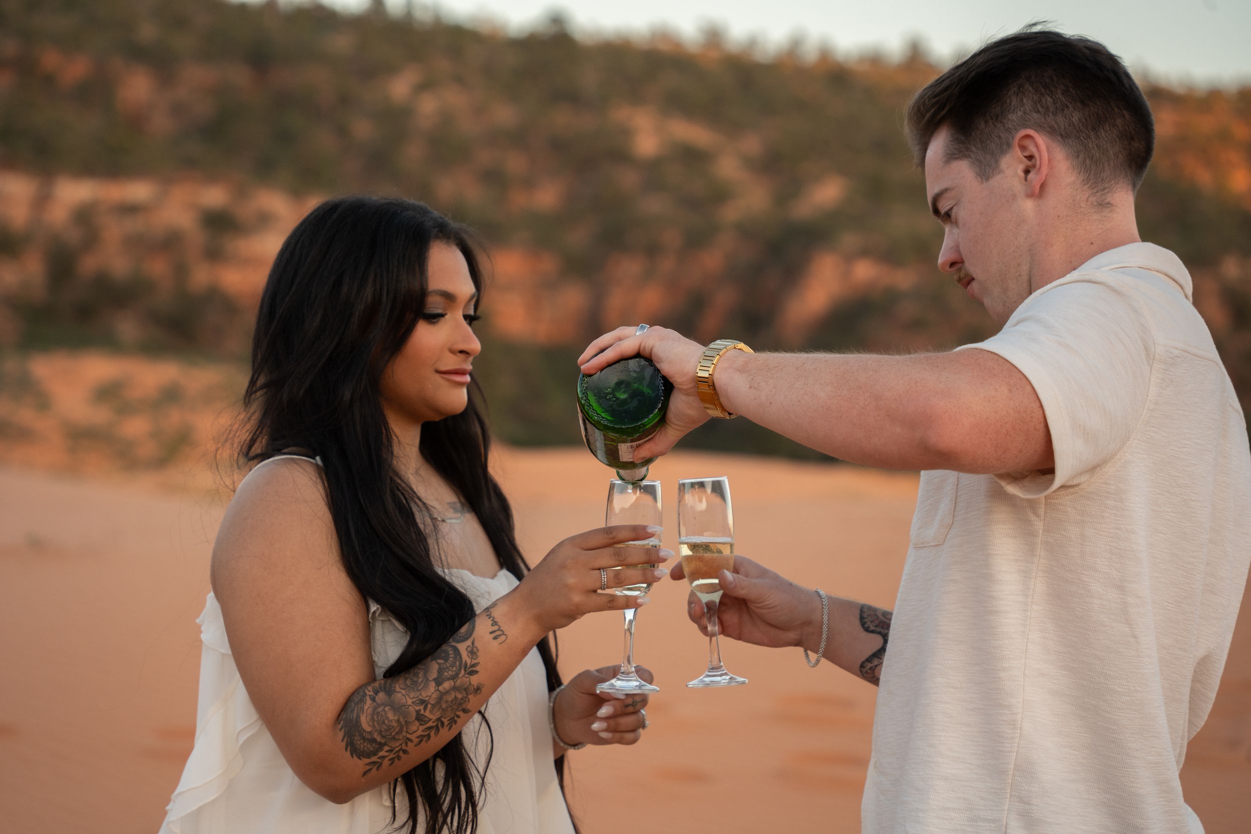 Couple celebrating proposal outdoors in desert, man pouring champagne into glasses at sunset.