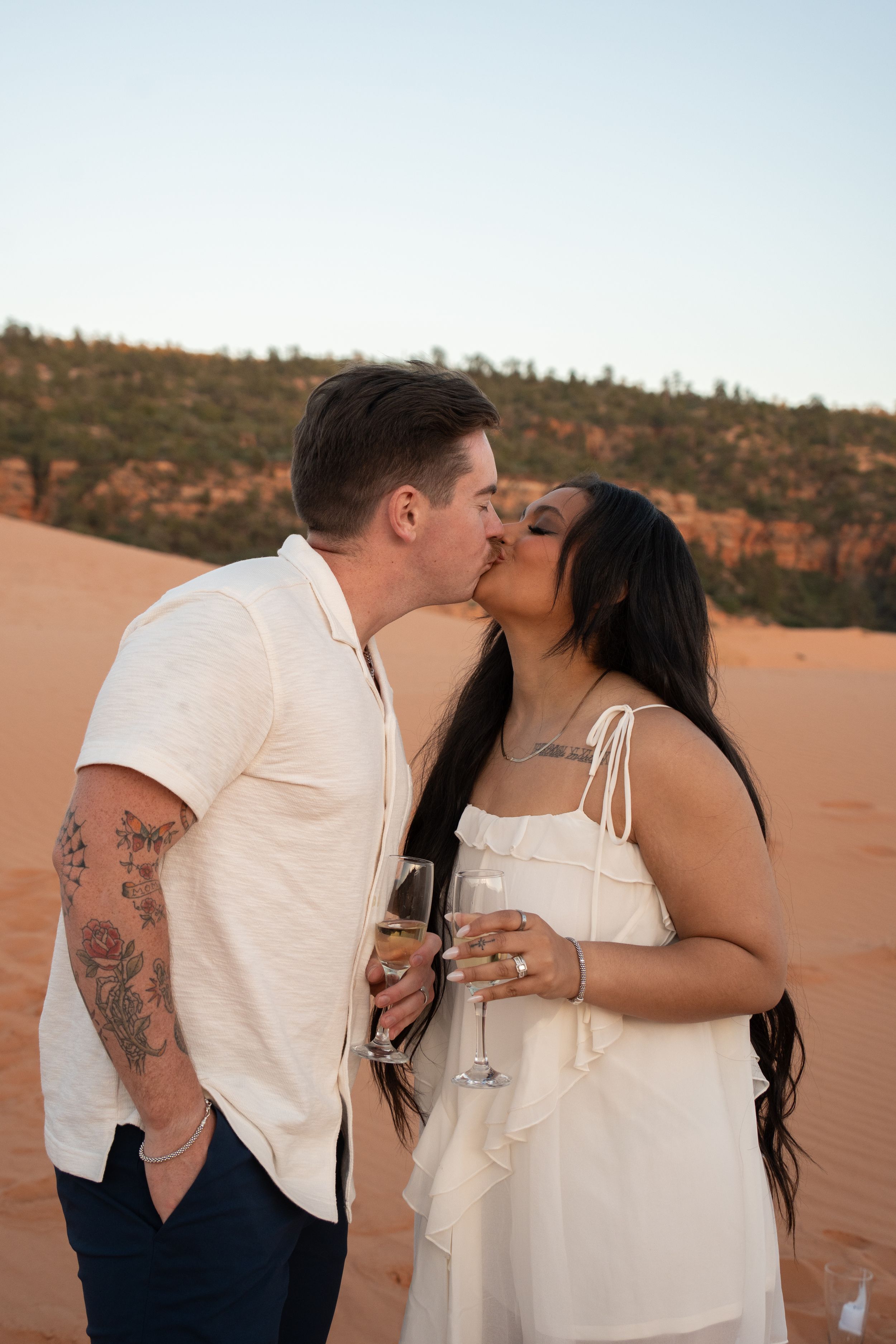 Couple kissing while holding champagne glasses and displaying engagement rings on red sand dunes at sunset, man with tattoos in white shirt.
