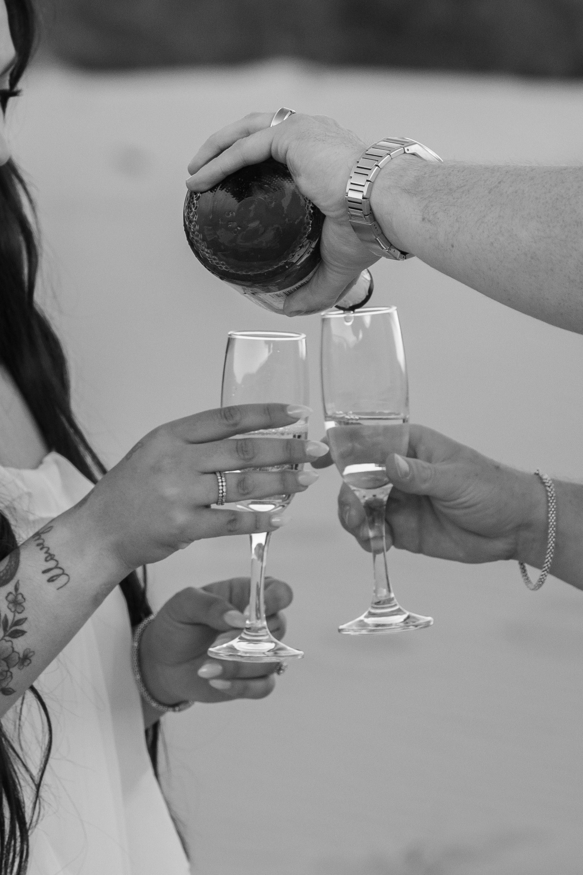 Black and white photo of hands holding champagne flutes while being filled from a bottle at a proposal celebration.