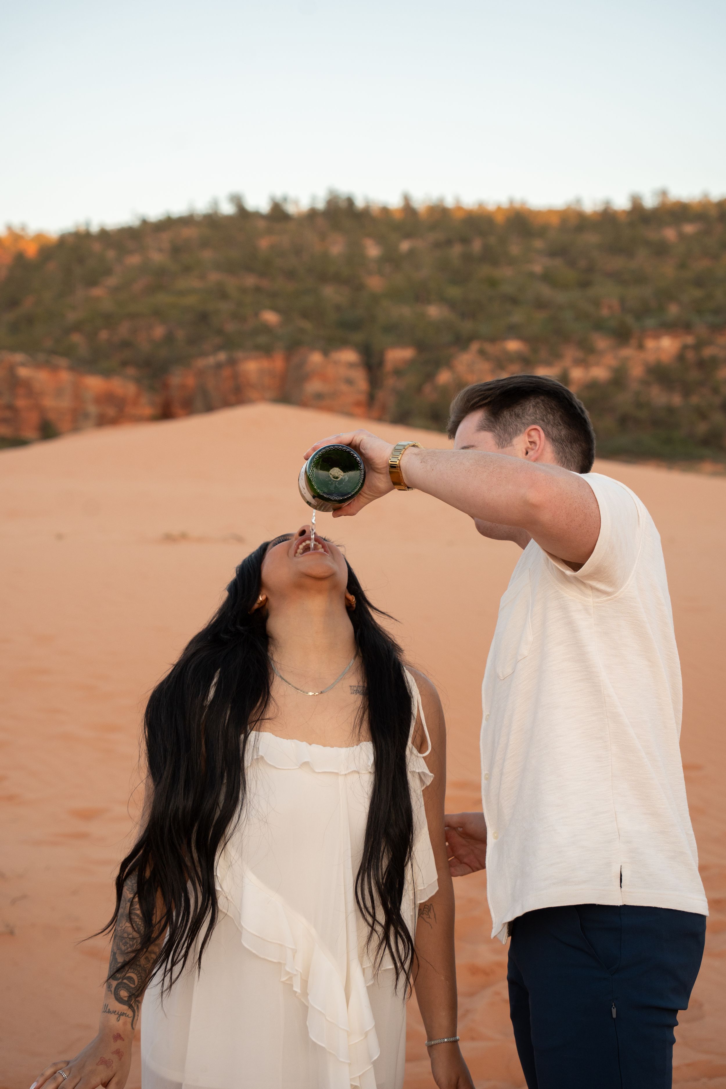 A man pours champagne from a green bottle into a woman's mouth on red sand dunes at sunset.