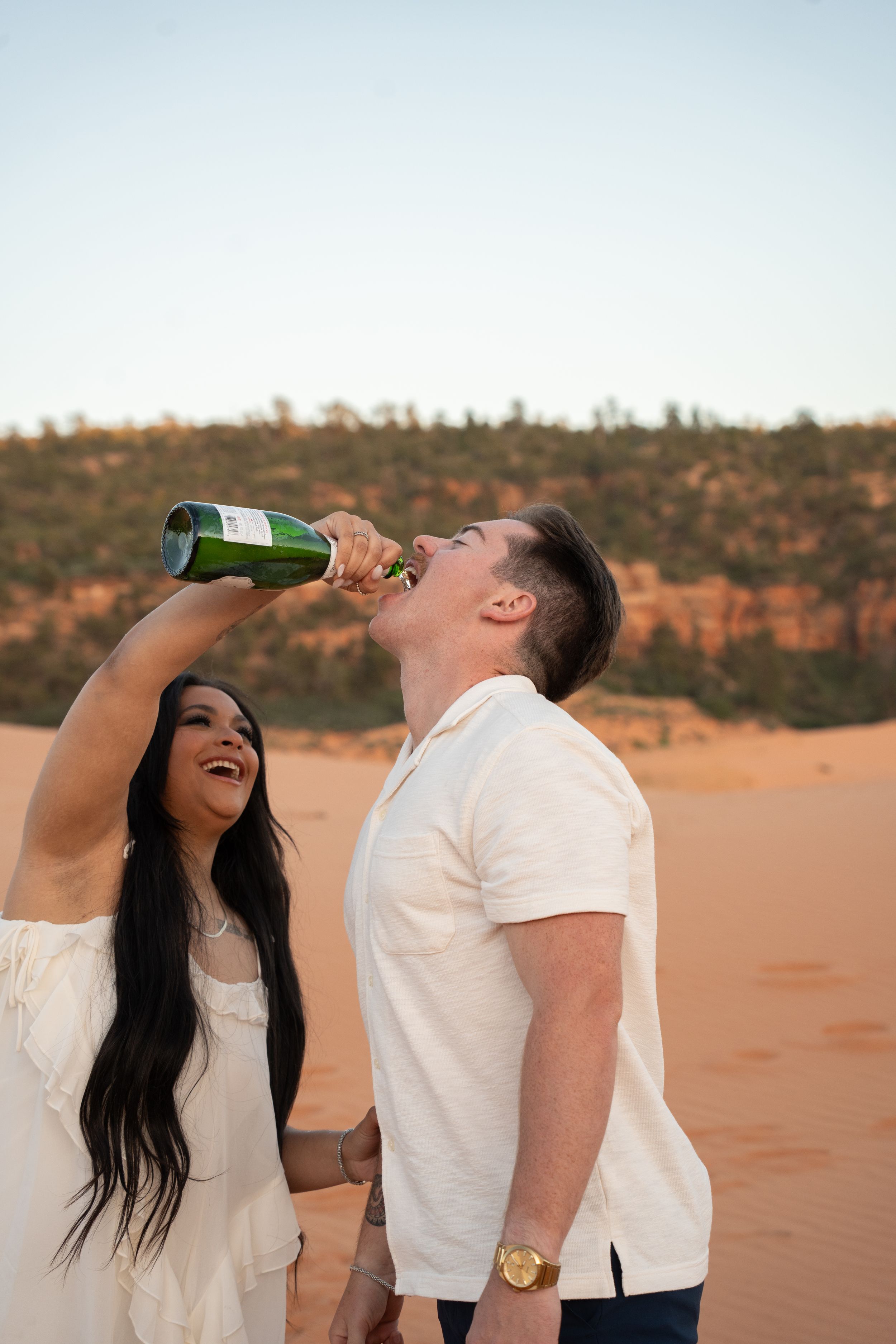 A couple in white outfits celebrates on red sand dunes, with the man drinking champagne from a green bottle.