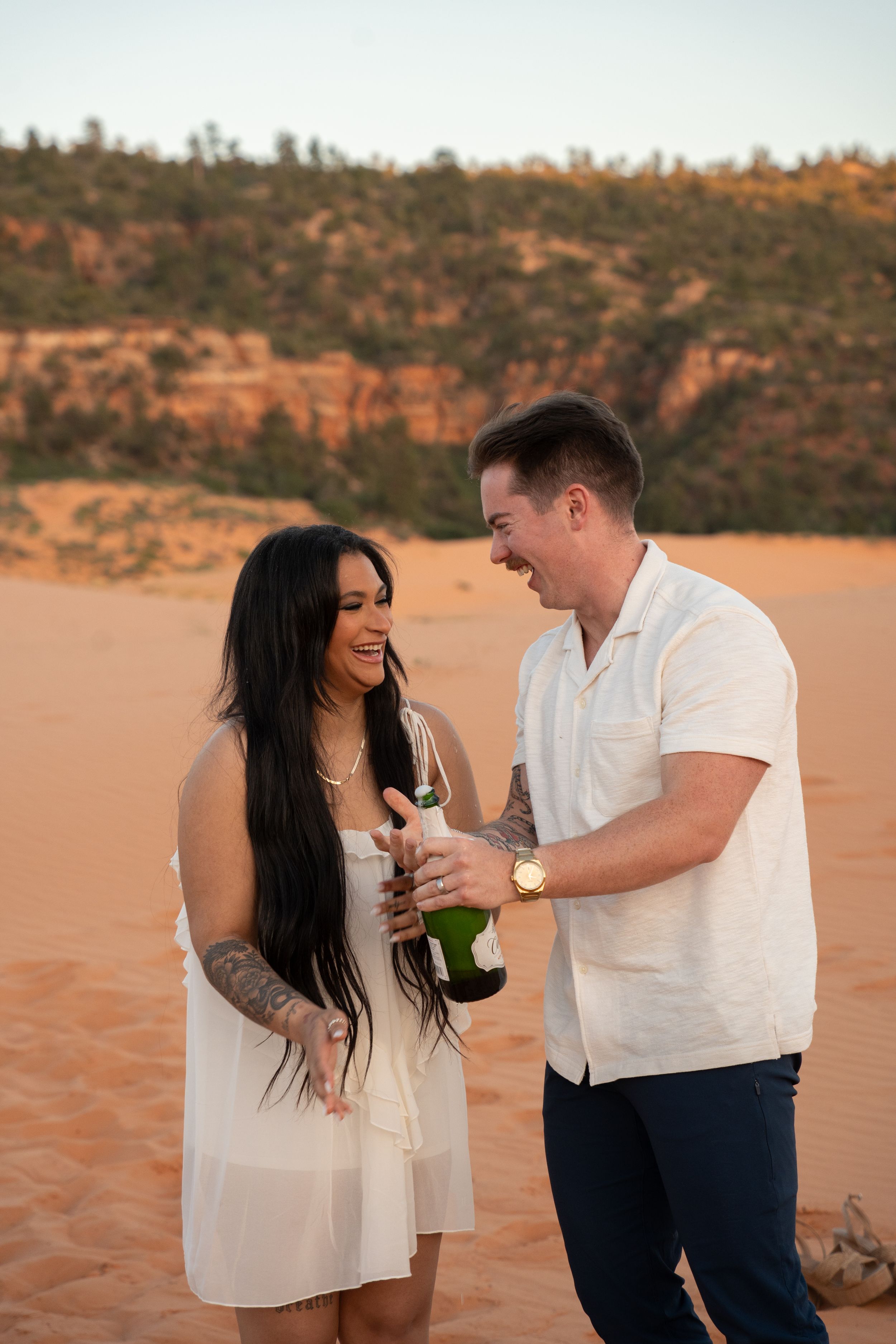 Couple celebrating on red sand dunes, opening champagne bottle, wearing white outfits, with red rock cliffs at sunset.