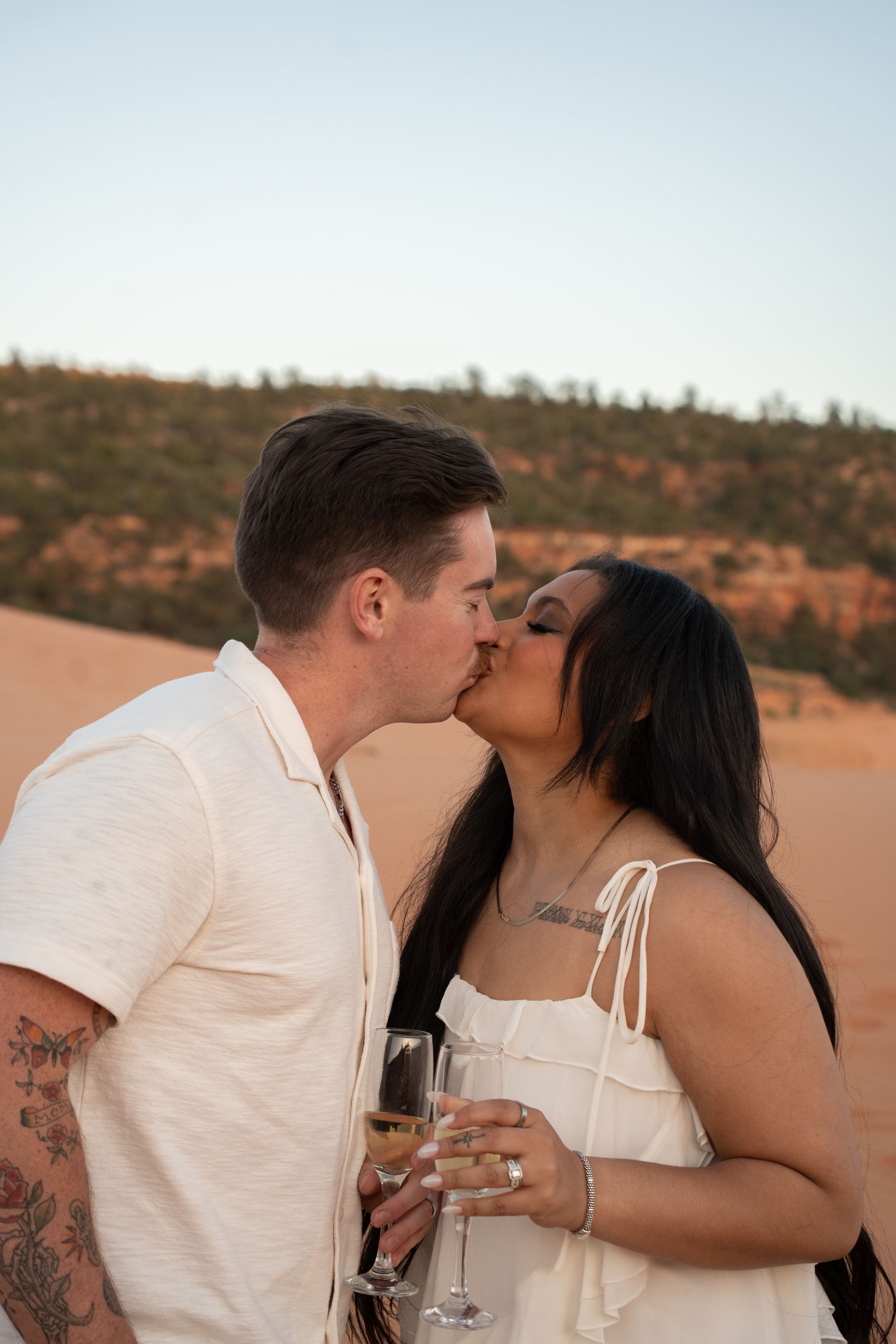 engaged Couple sharing a romantic kiss while holding champagne glasses on coral pink sand dunes at sunset.
