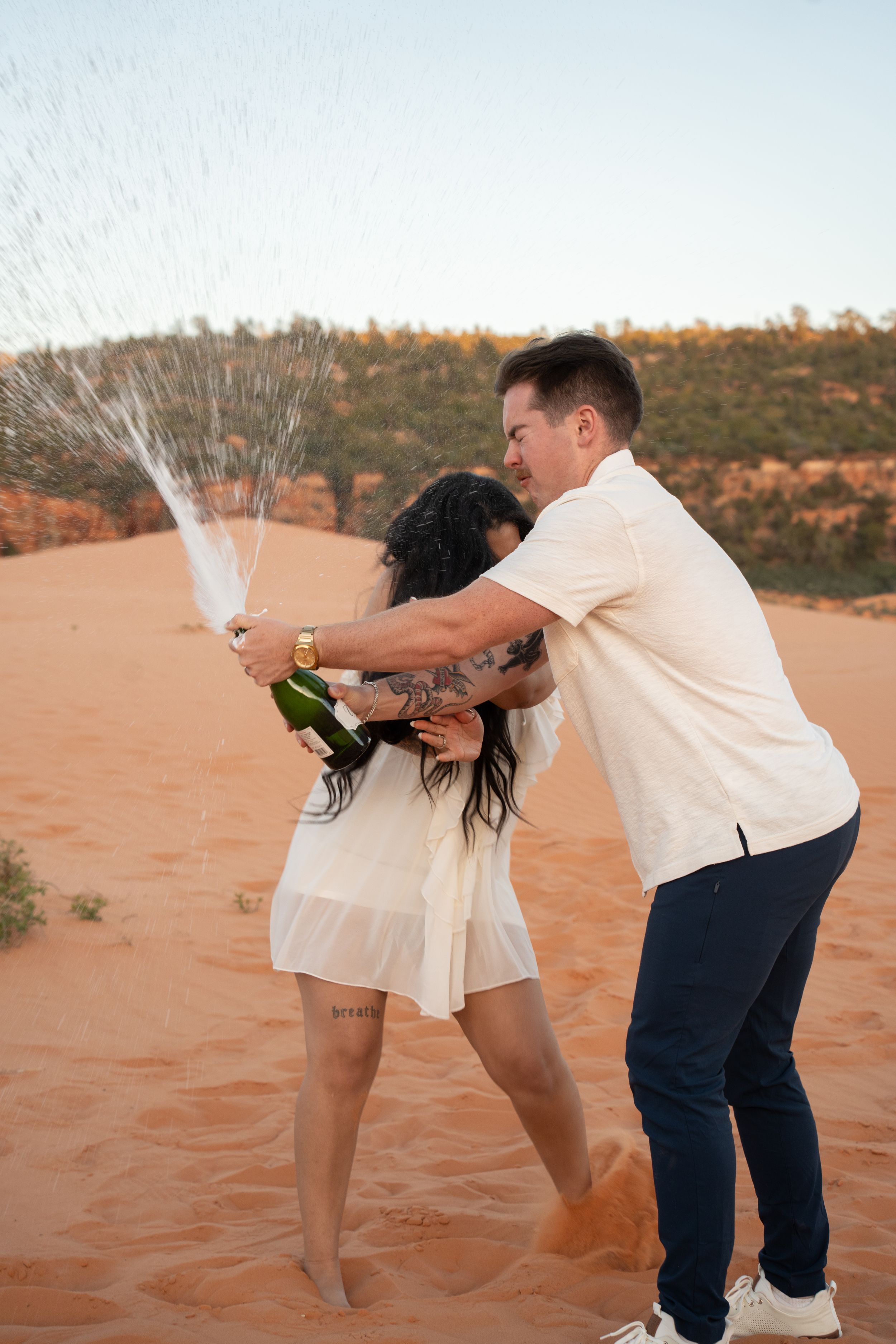 Couple celebrating on red sand dunes by popping champagne, woman in white dress, man in white shirt and navy pants.