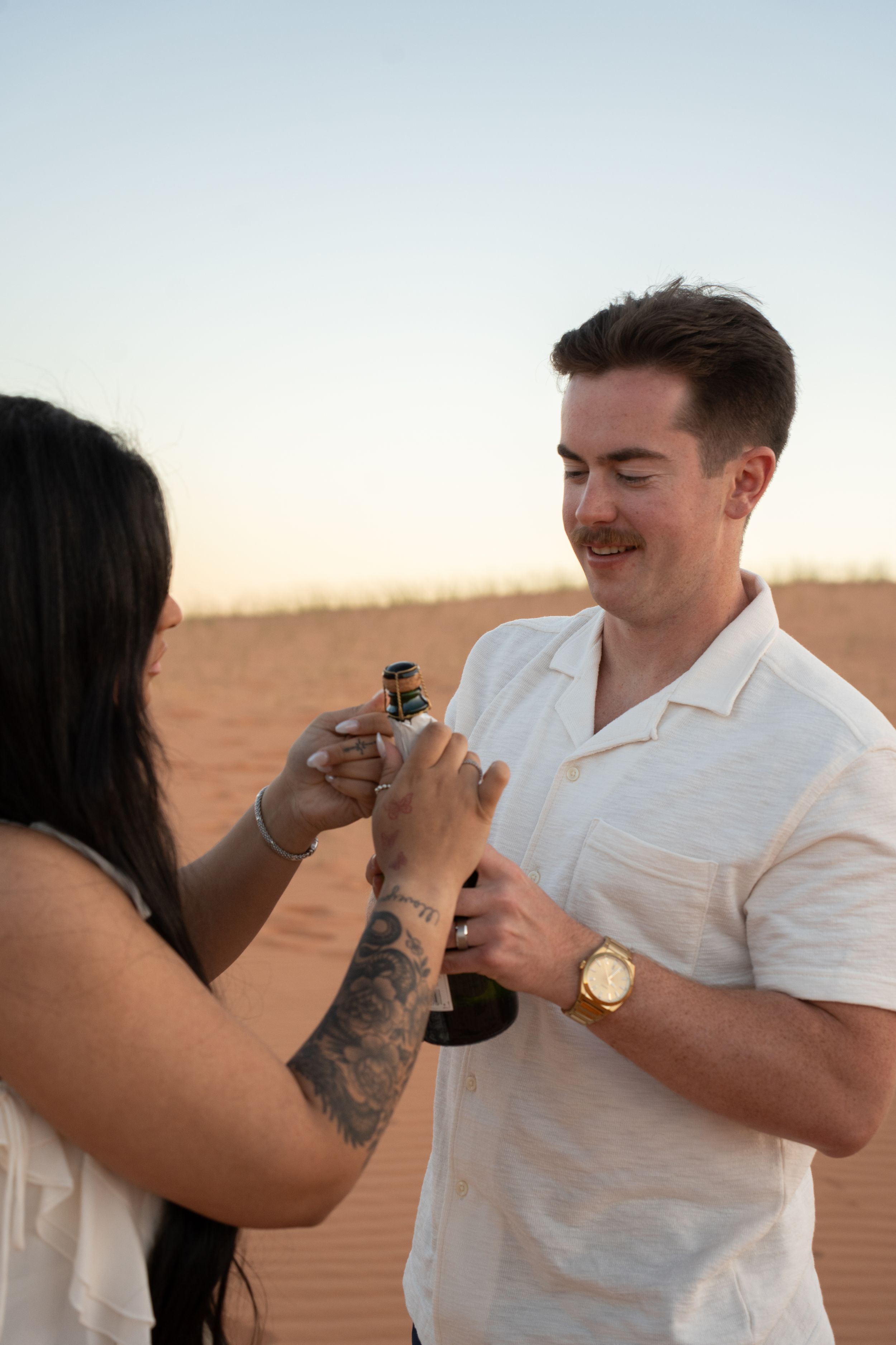 A man and woman opening a champagne bottle together in a red dune landscape at sunset.