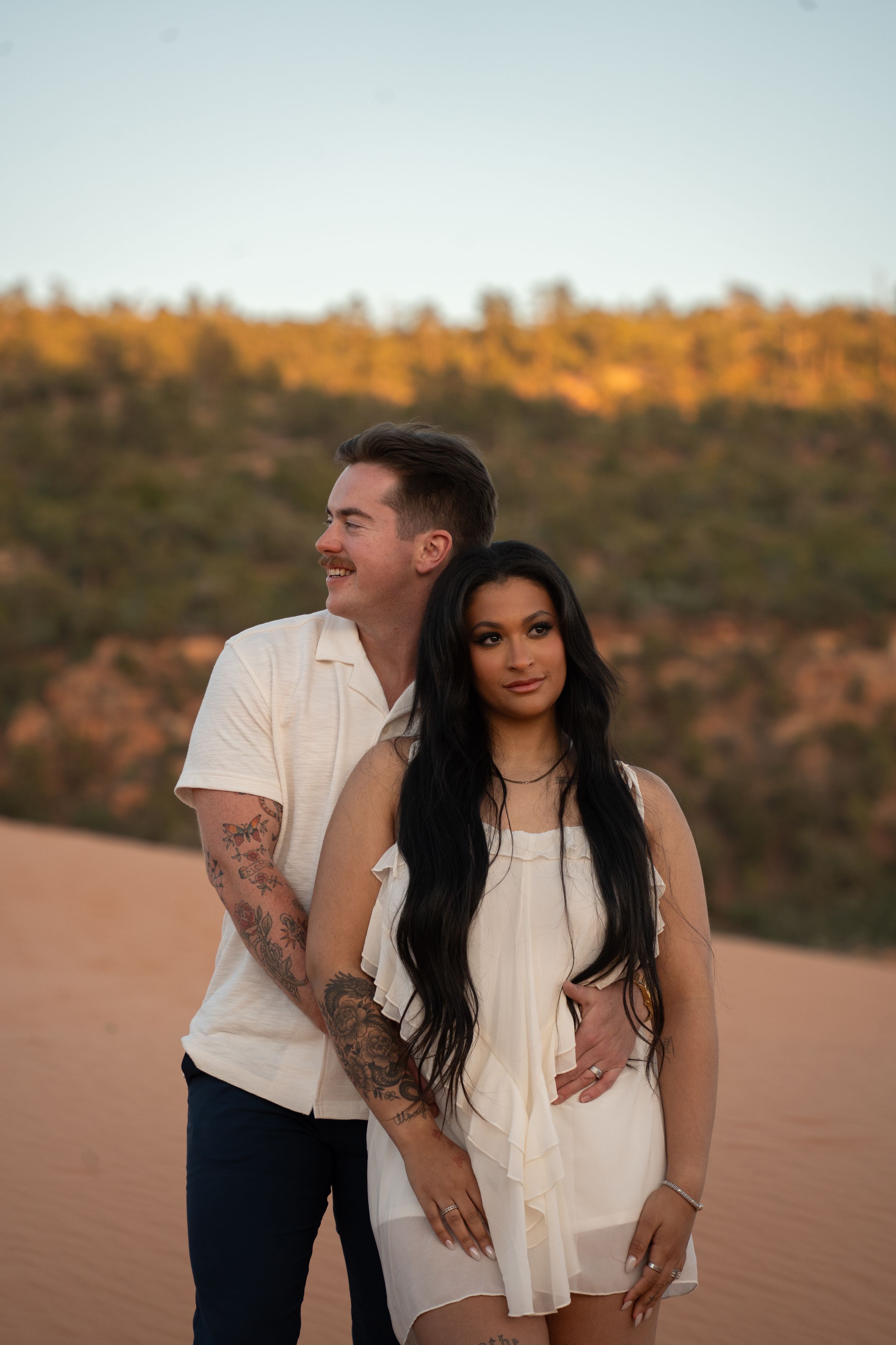 Couple in white outfits posing on sandy dunes at golden hour with lush greenery in the background.