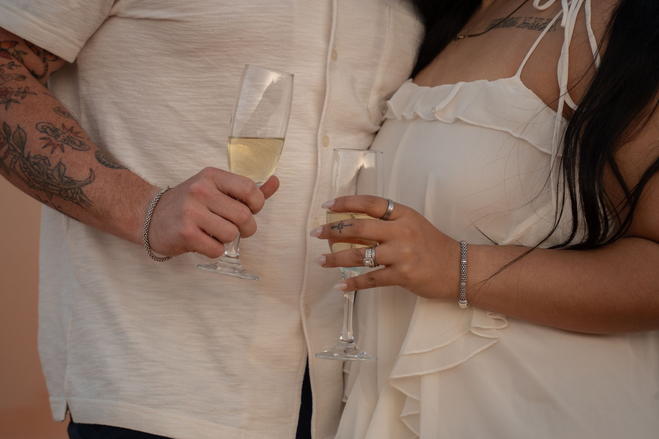 Couple in white outfits holding champagne flutes, man with tattoos, woman wearing engagement ring and bracelet.
