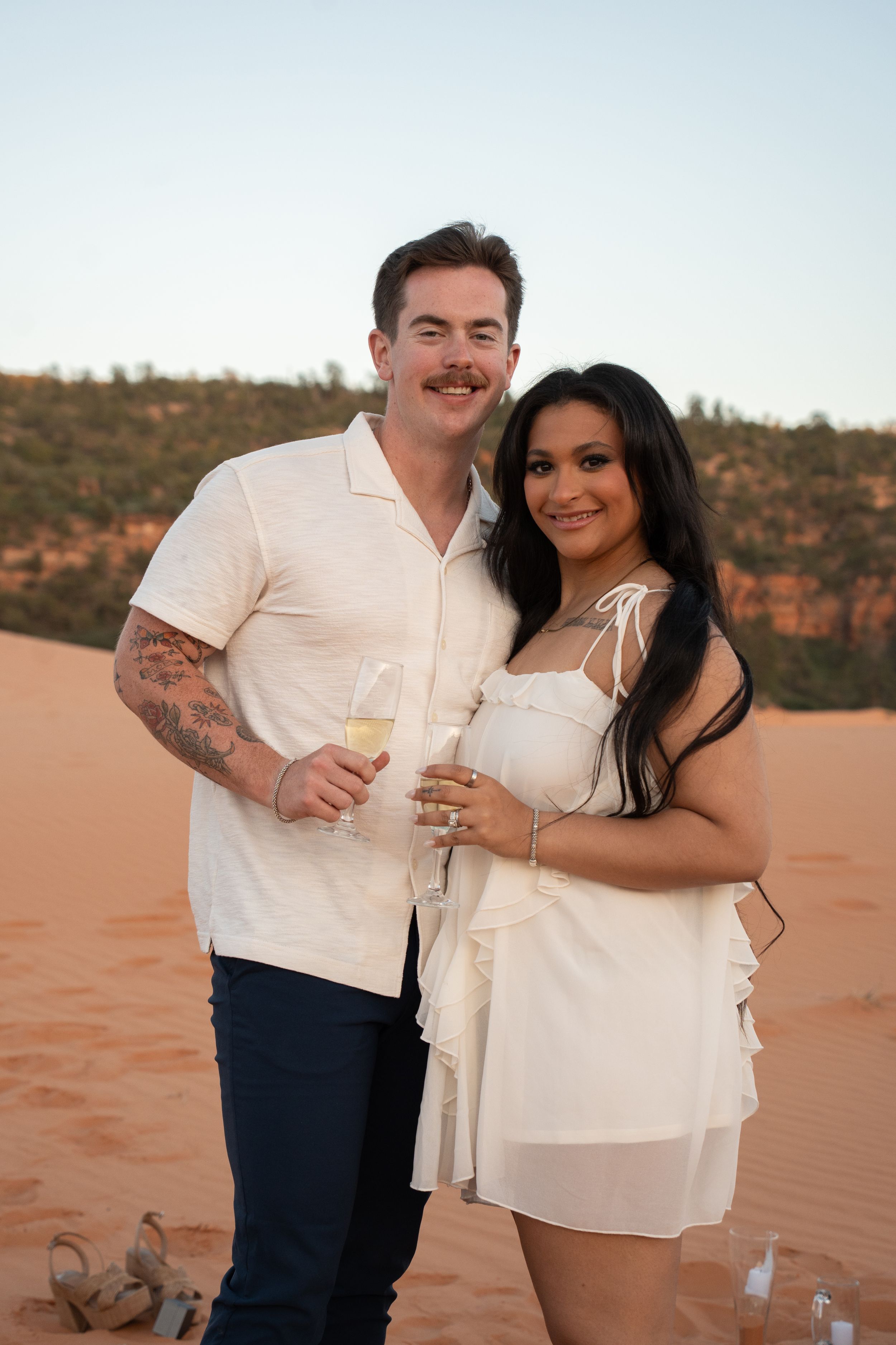 newly engaged Couple in white outfits holding champagne glasses on red sand dunes at sunset.