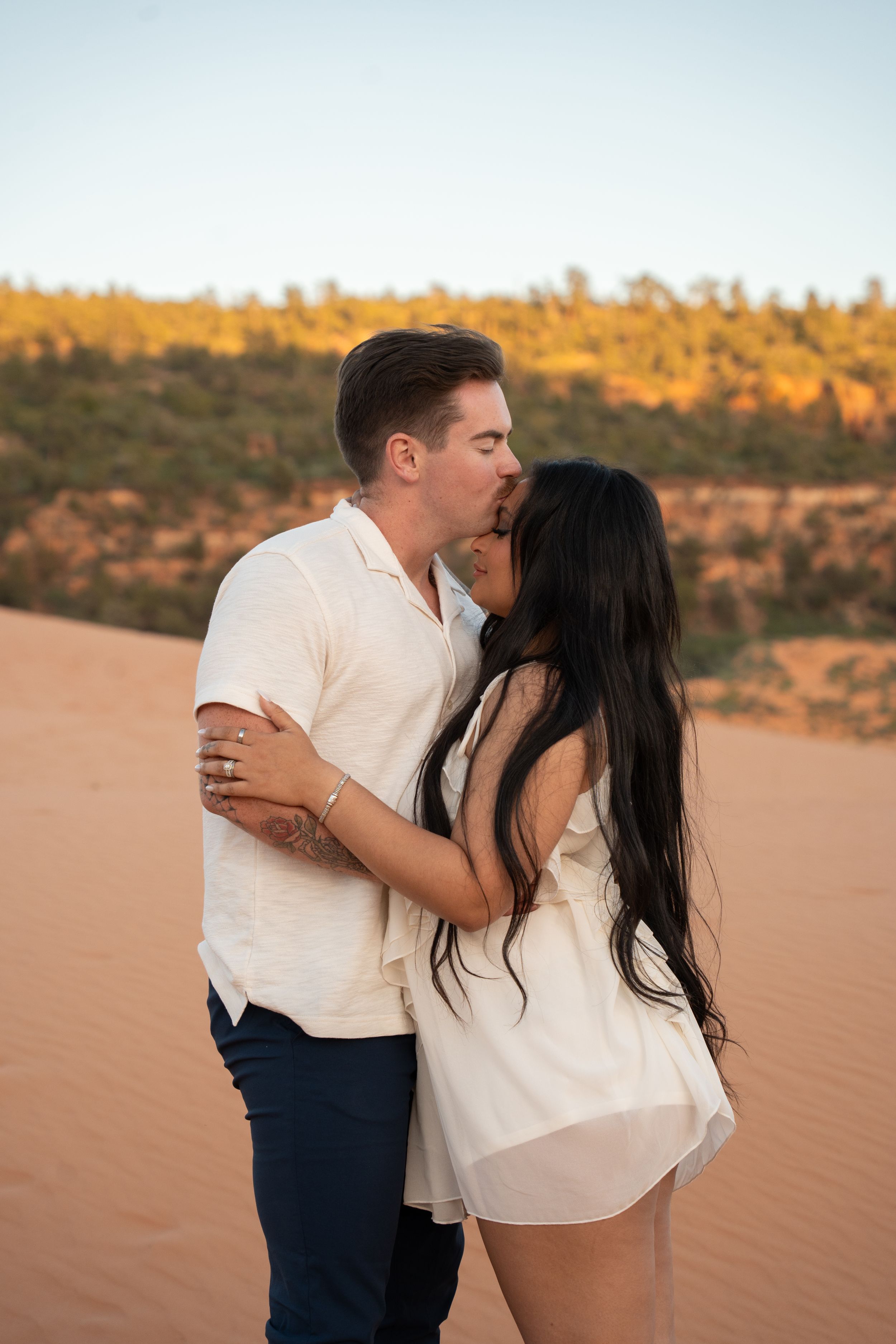 Couple sharing a romantic kiss on pink sand desert dunes at golden hour, woman in white dress, man in white shirt.