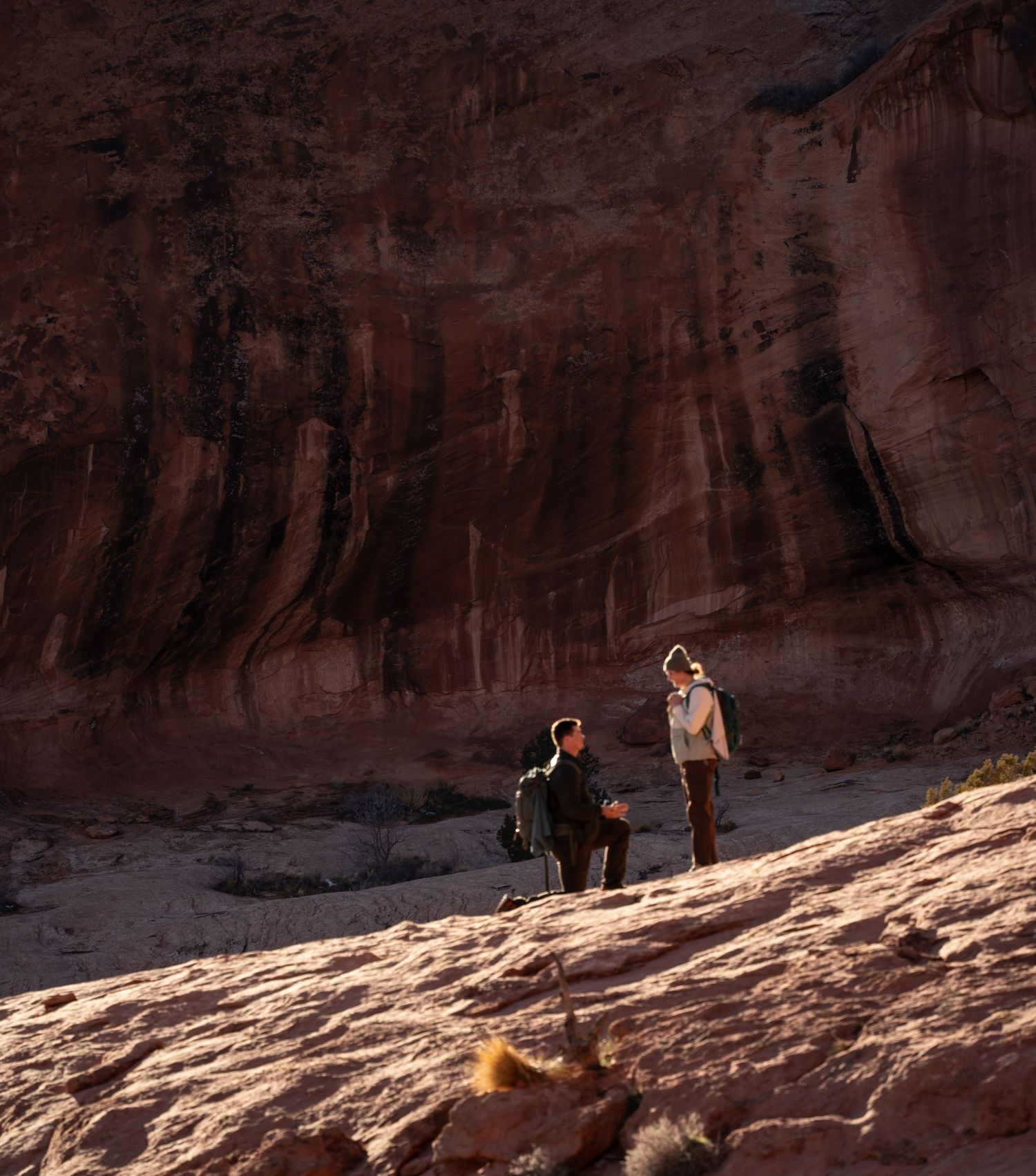 Two hikers stand on red rock formations before a towering desert arch