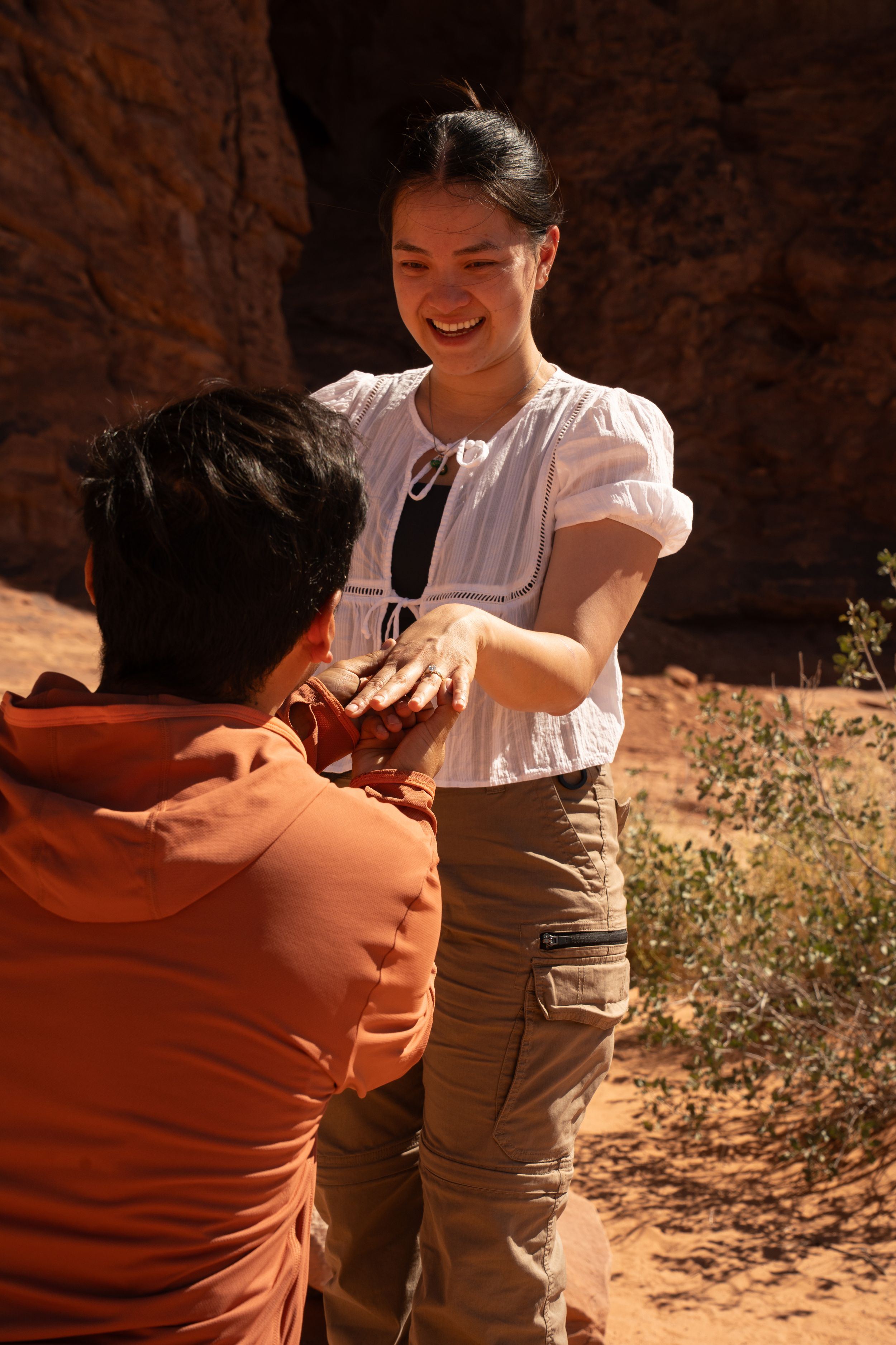 Man kneeling proposing to smiling woman in white blouse and khaki pants at double arch in arches national park