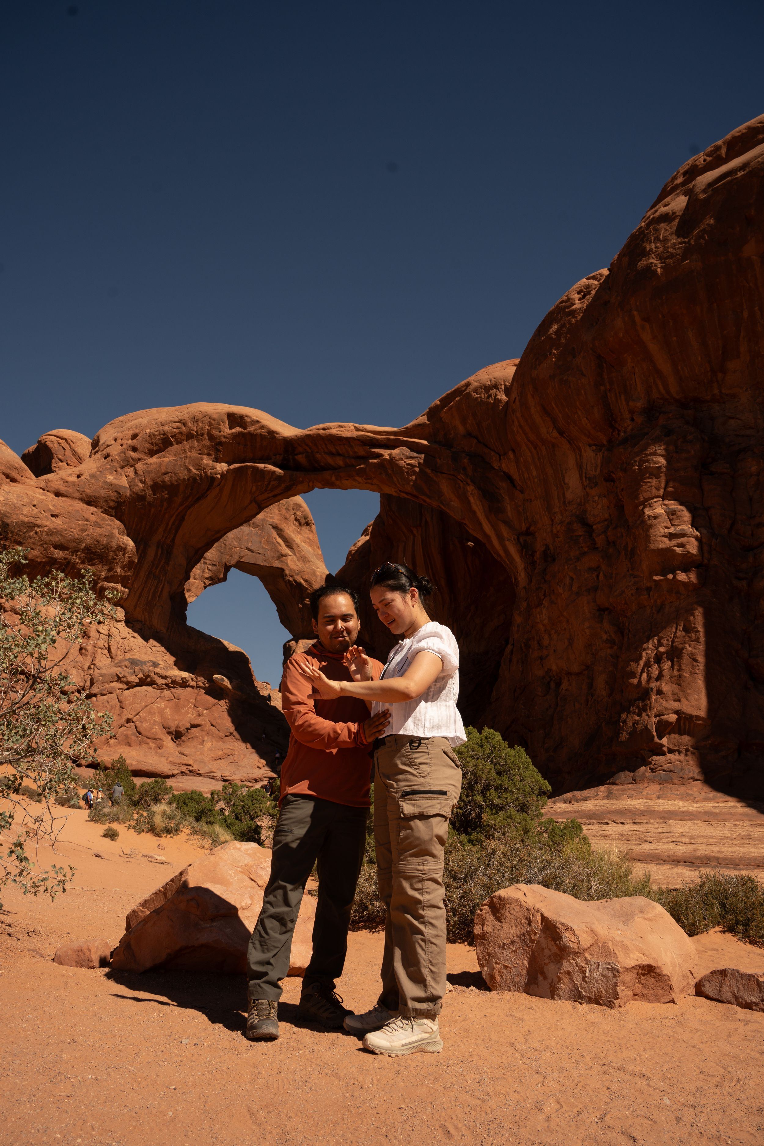 Two people embrace before a stunning red sandstone arch formation under a deep blue sky in a desert landscape.