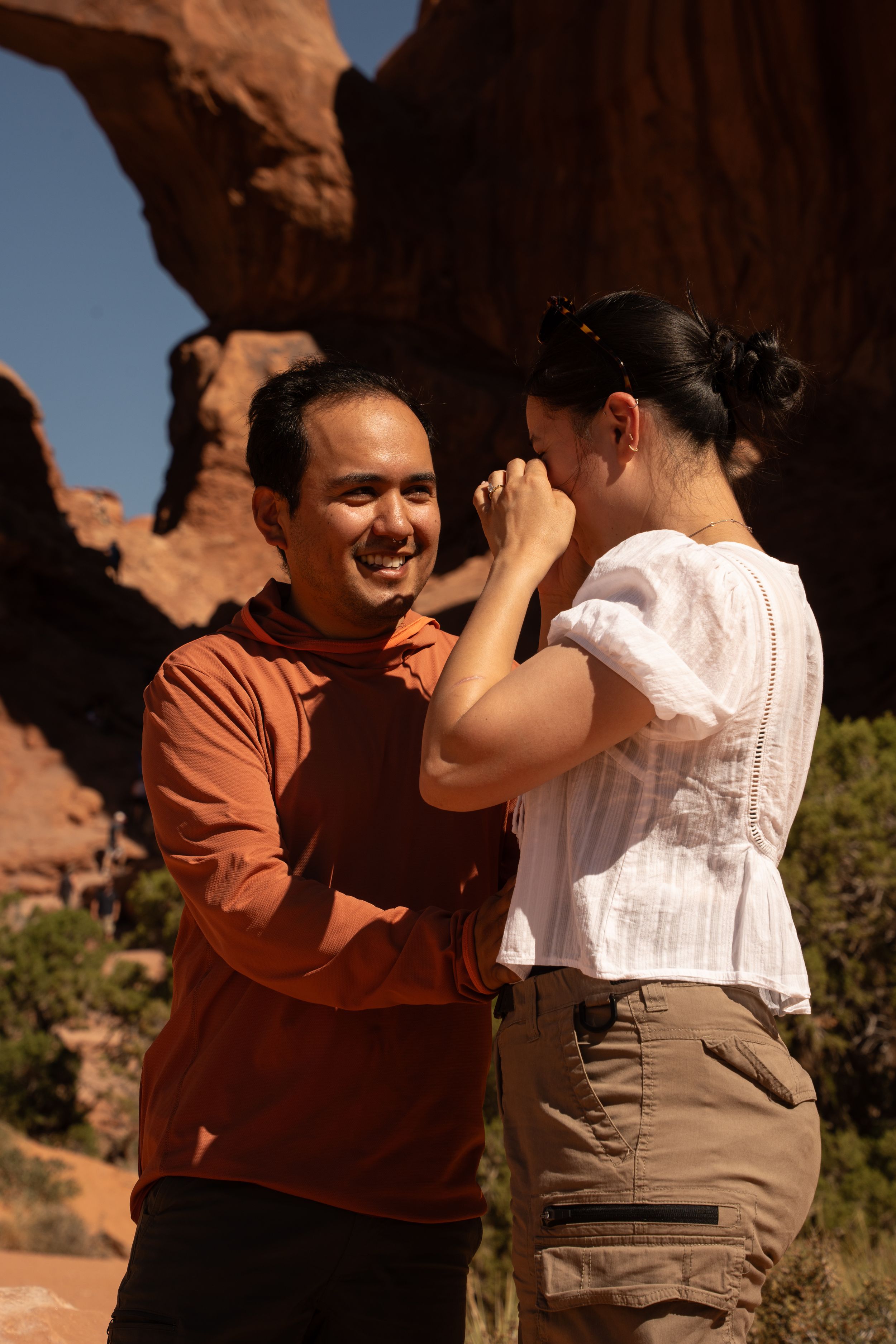 Couple sharing a romantic moment at Arches National Park, man in orange hoodie smiling at woman in white top.