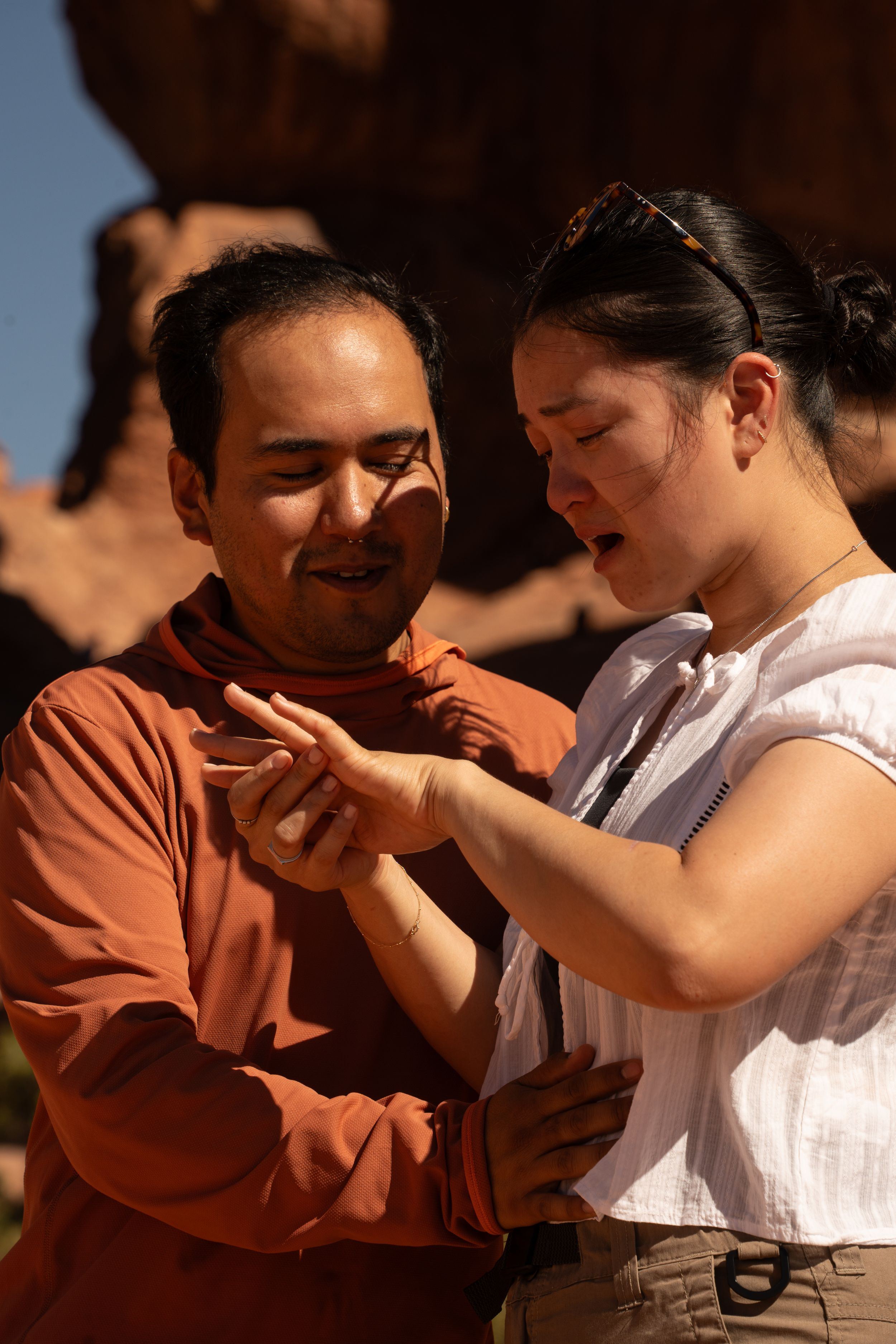 A man and woman examine her engagement ring outdoors near red rock formations in natural sunlight.