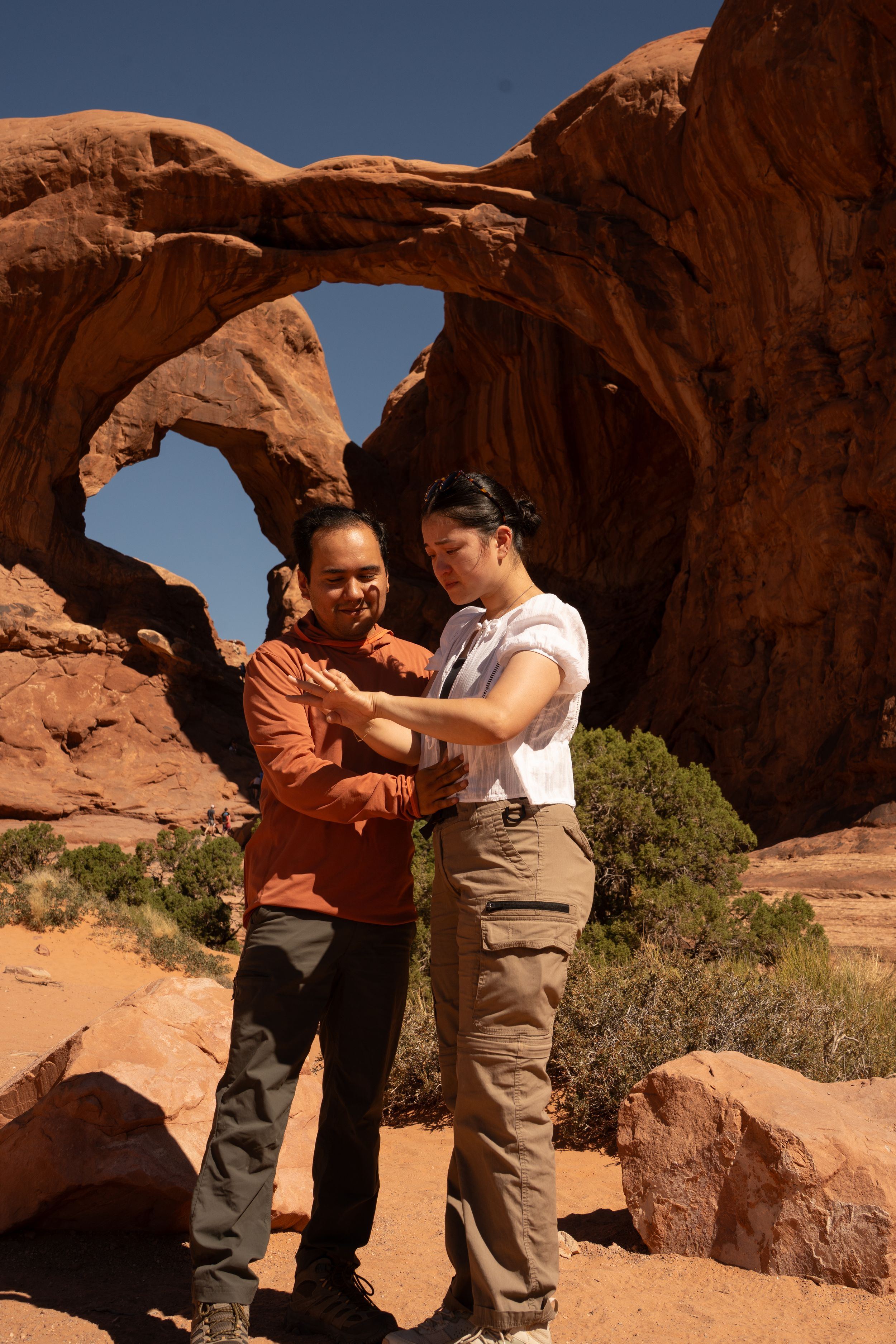 Two people stand together before a stunning natural rock arch formation in a desert landscape under a clear blue sky.