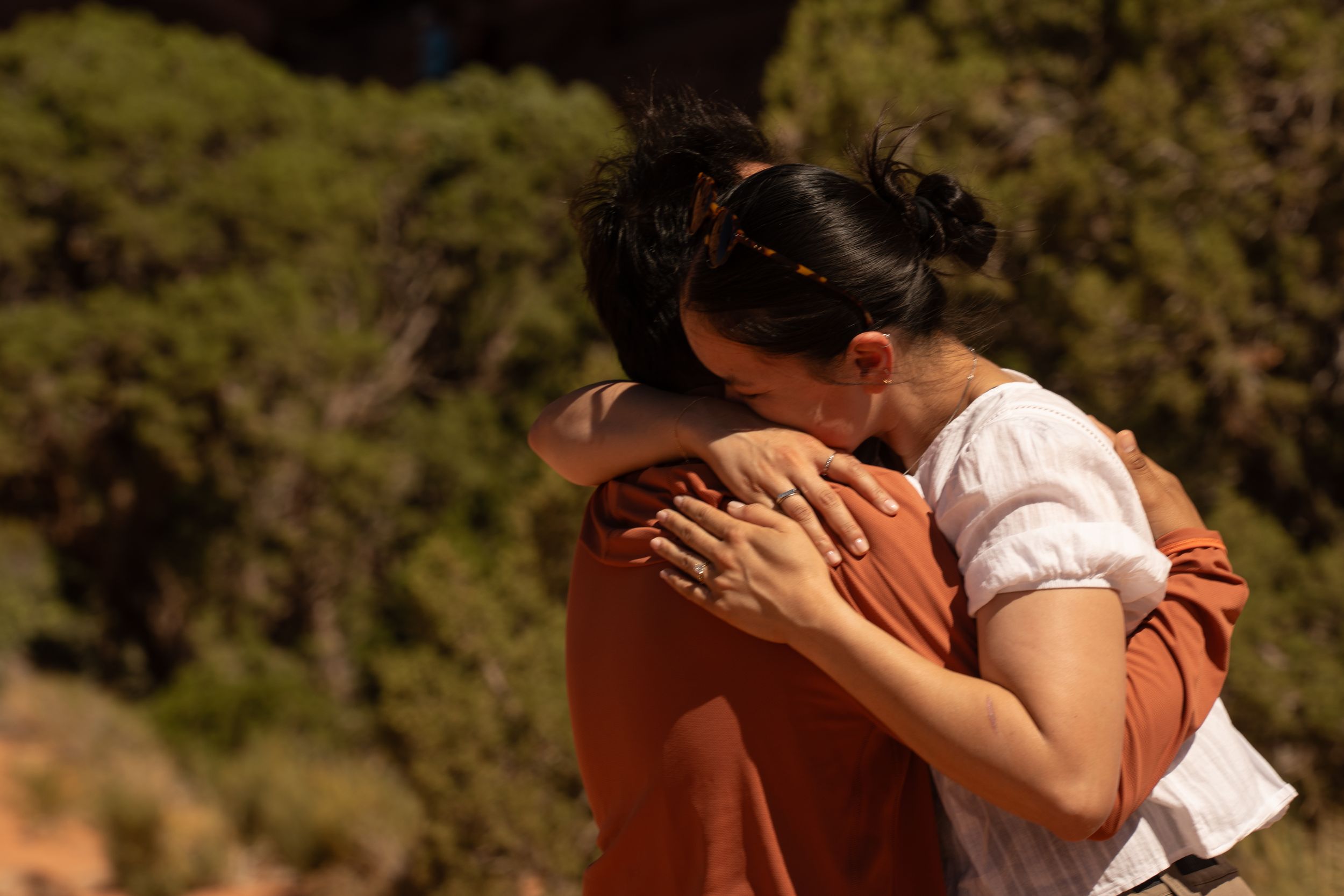 Two people sharing a warm embrace outdoors, surrounded by green trees in natural sunlight.