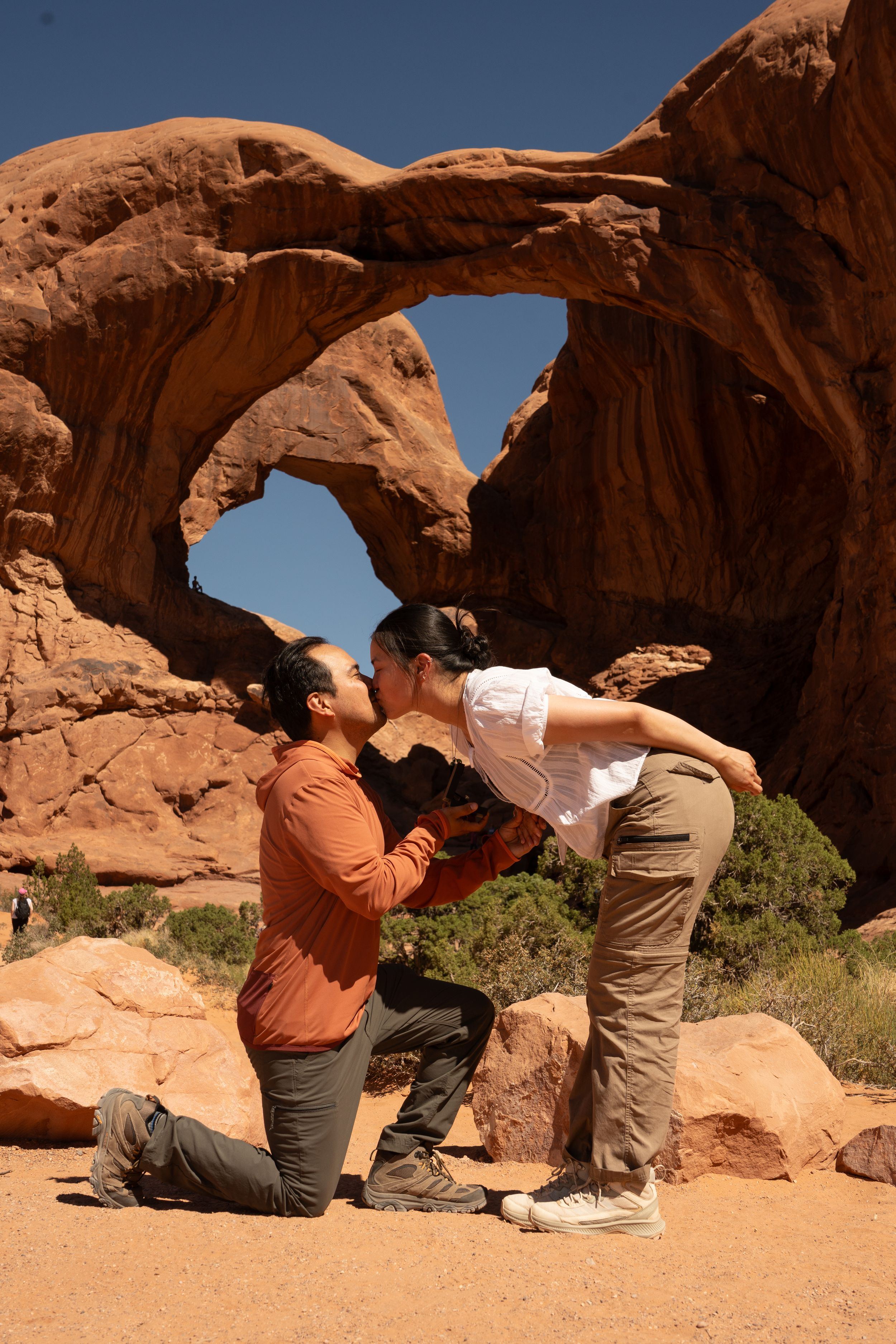 Couple sharing a romantic kiss on one knee at Double Arch in Arches National Park, Utah desert landscape.