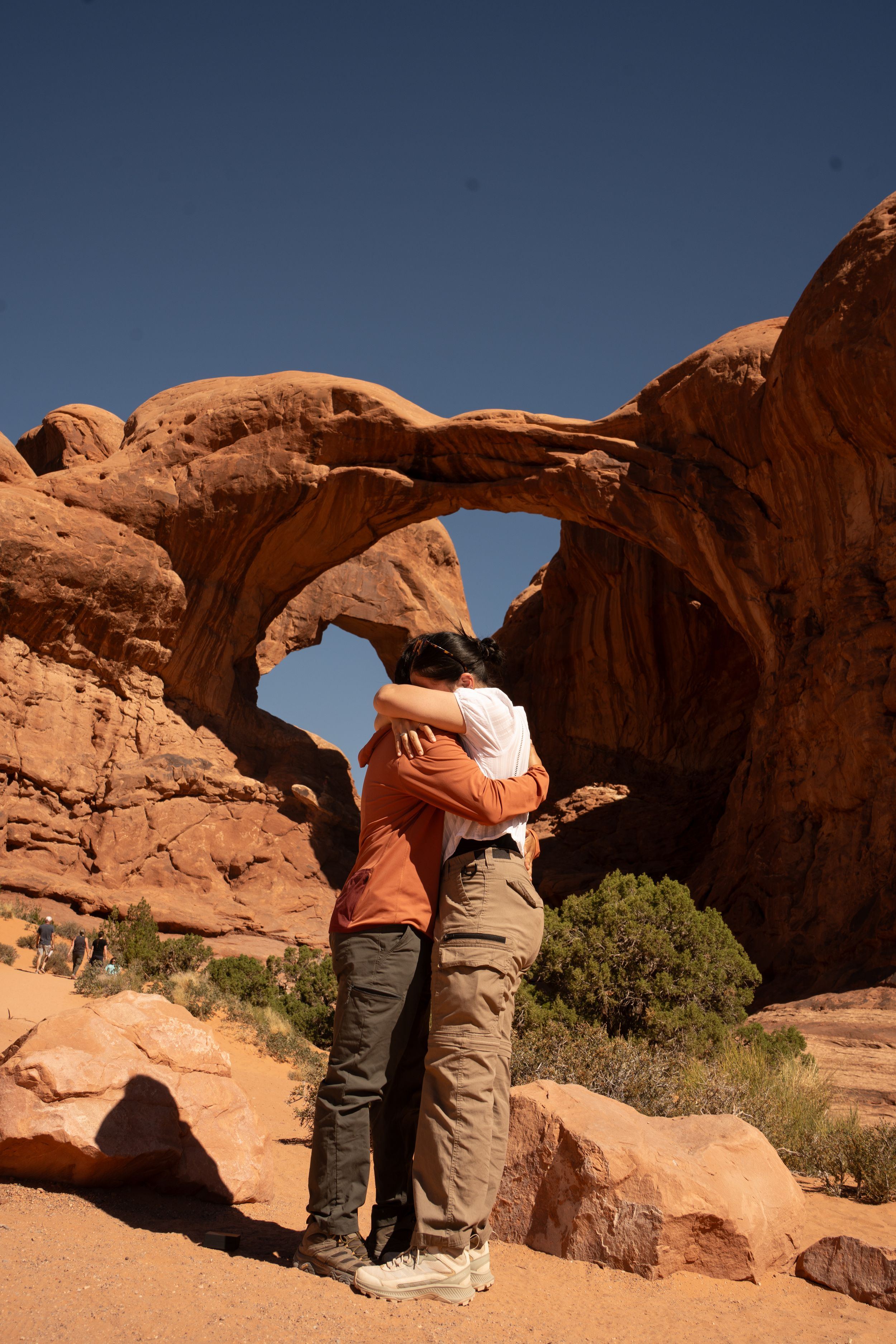 Couple embracing in front of a stunning red rock arch formation in Arches National Park, Utah desert landscape.