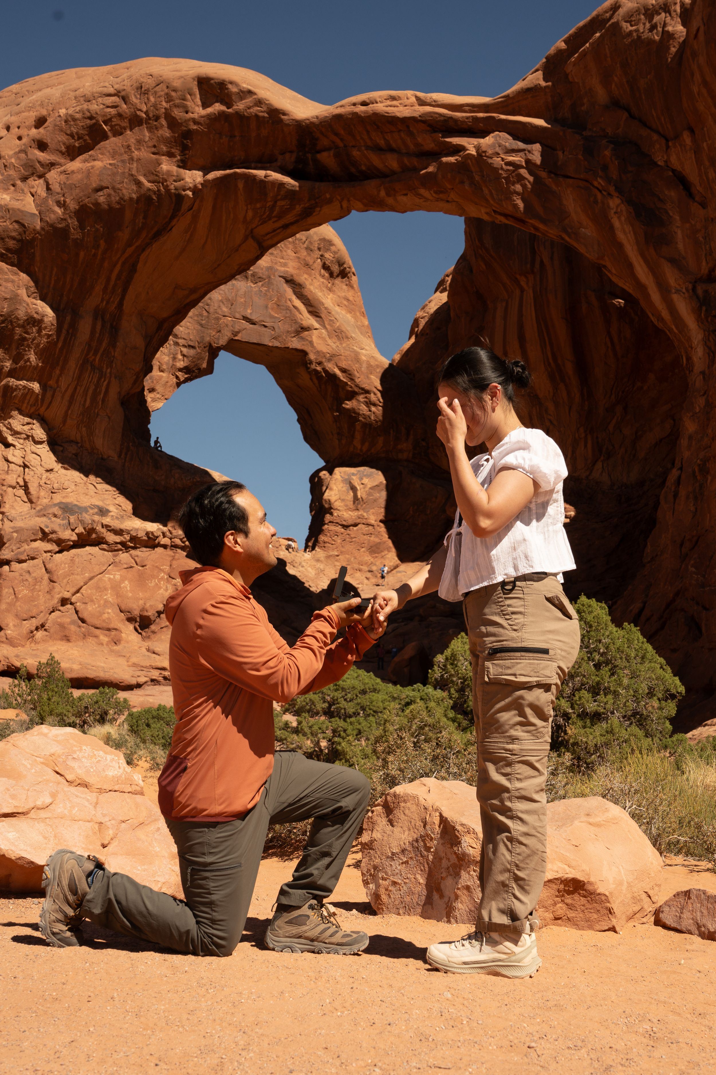 Man proposes on one knee to woman at Arches National Park, Utah, with iconic red rock arch formation behind them.