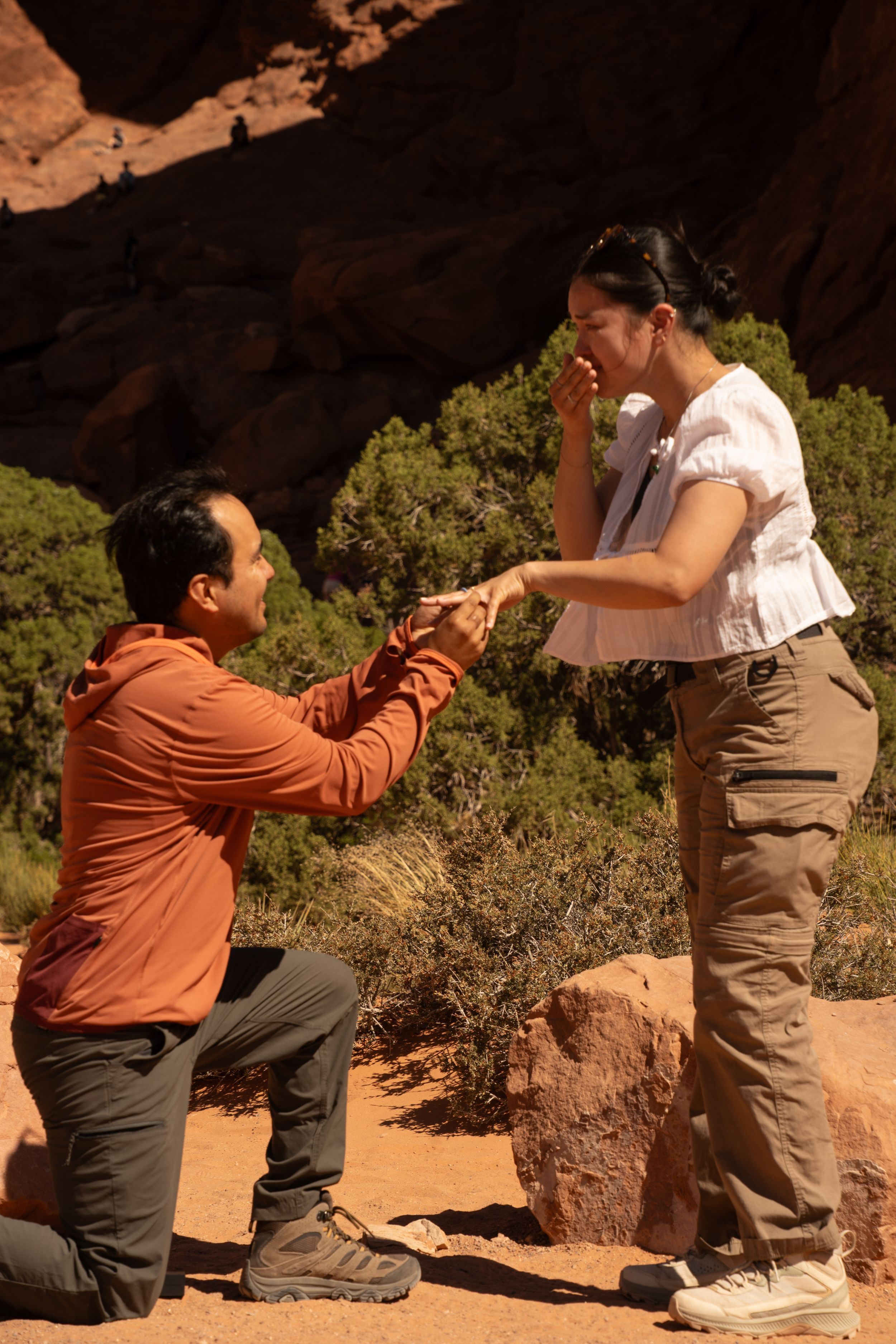 Man proposing to woman on one knee at red rocks in moab utah, she covers her mouth in surprise.