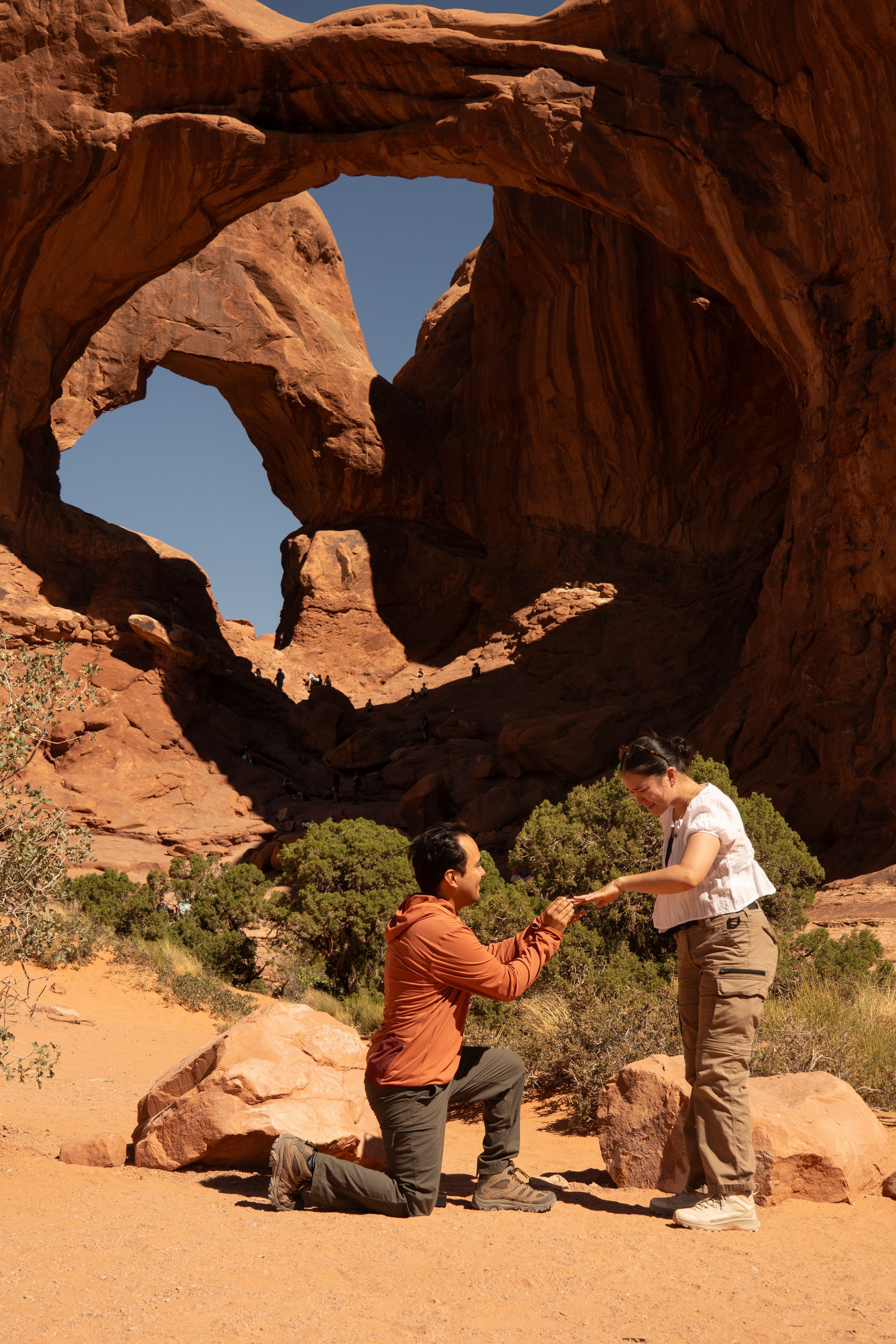 Person proposing on one knee at Double Arch in Arches National Park, Utah, with red sandstone formations backdrop.