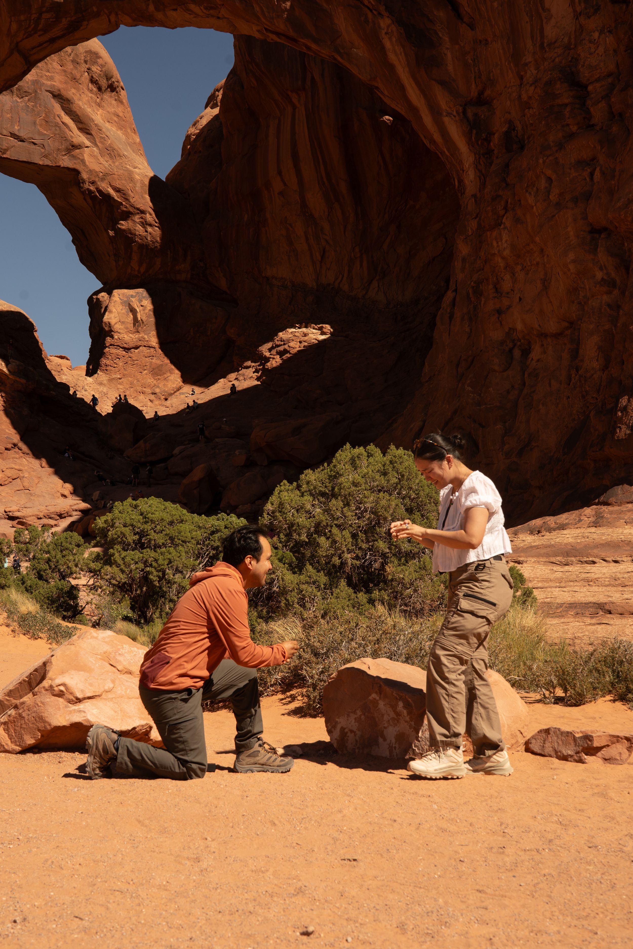 Man proposing to woman on one knee beneath a dramatic red rock arch in Arches National Park, Utah.