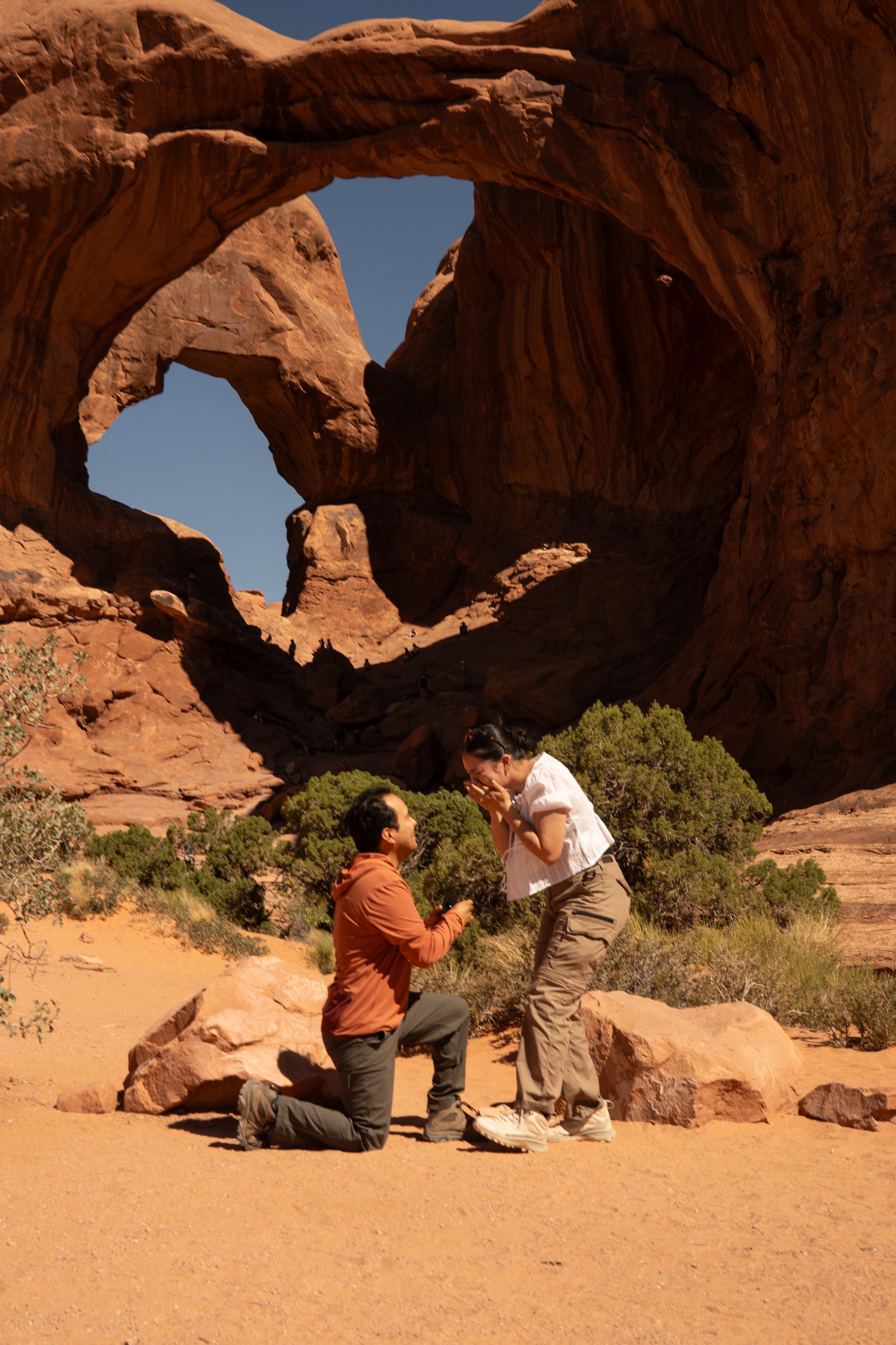 Person proposing on one knee at Double Arch in Arches National Park, Utah, with red rock formations under blue sky.