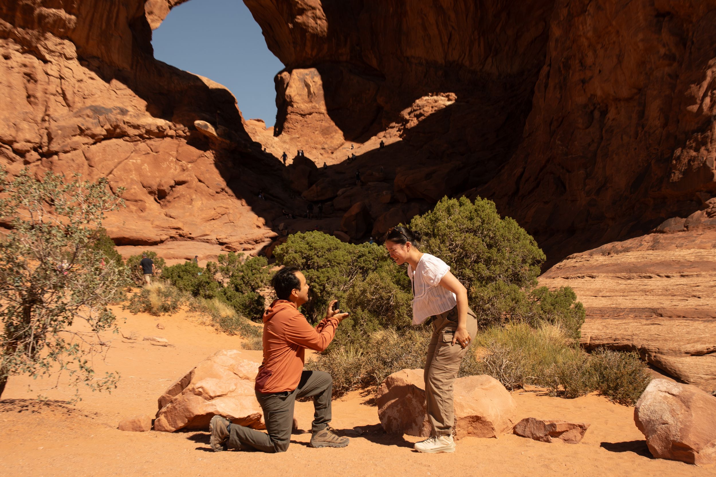 Person proposing on one knee to partner at Arches National Park with stunning red rock arch formation in background.