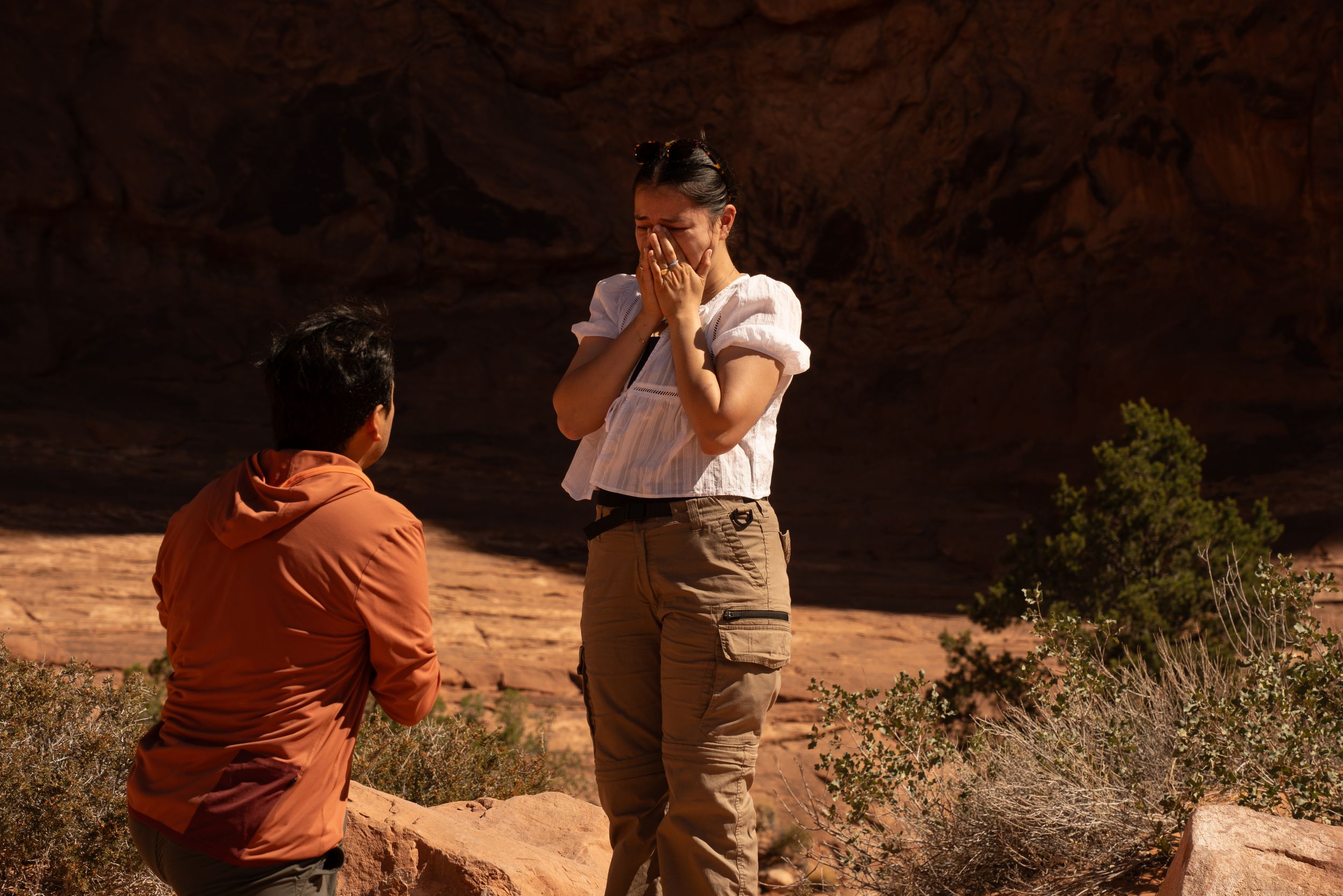 Man kneeling proposing to surprised woman covering her face in red rock canyon desert landscape.
