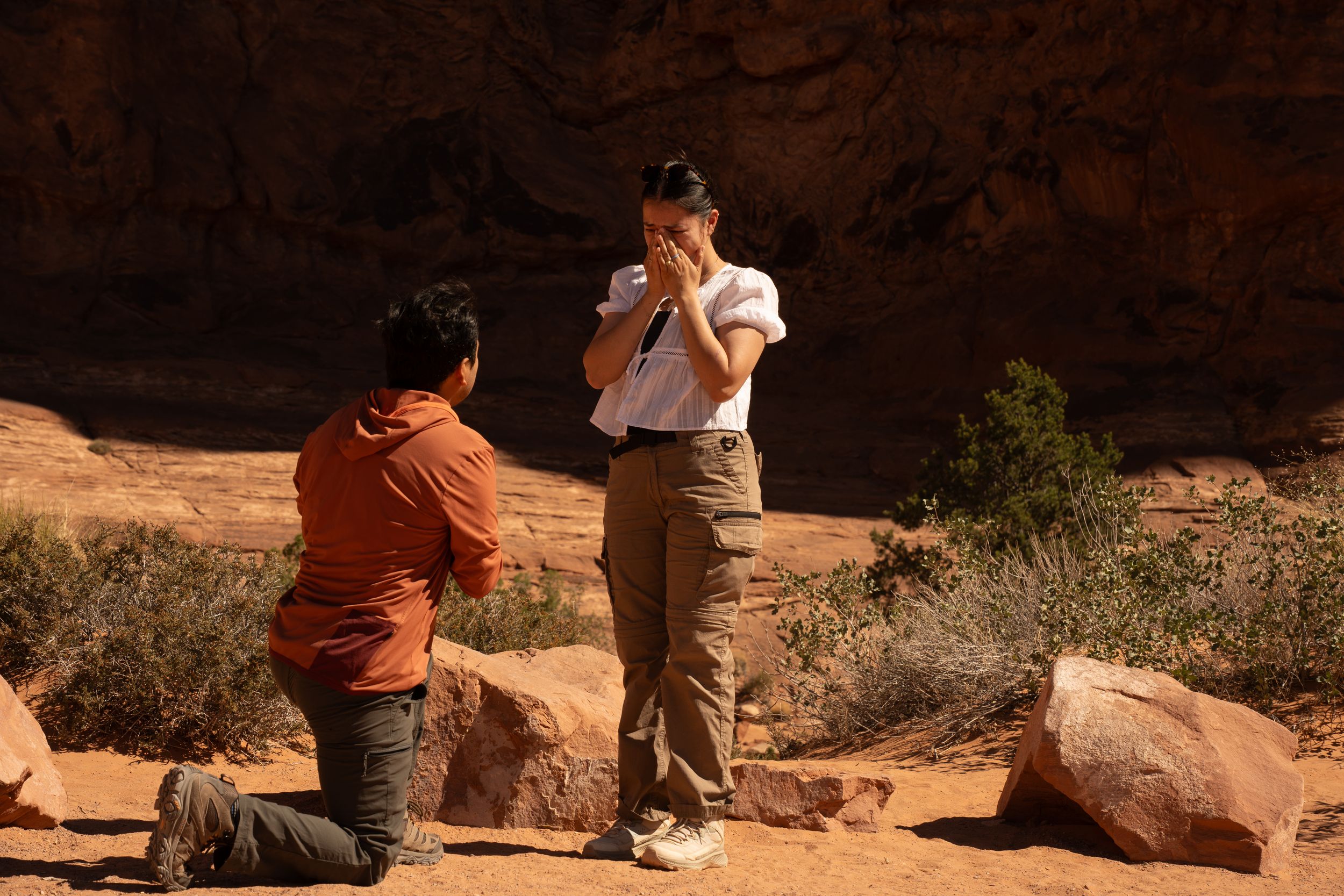 Person proposing on one knee to a surprised partner among red rock formations in a desert canyon setting.