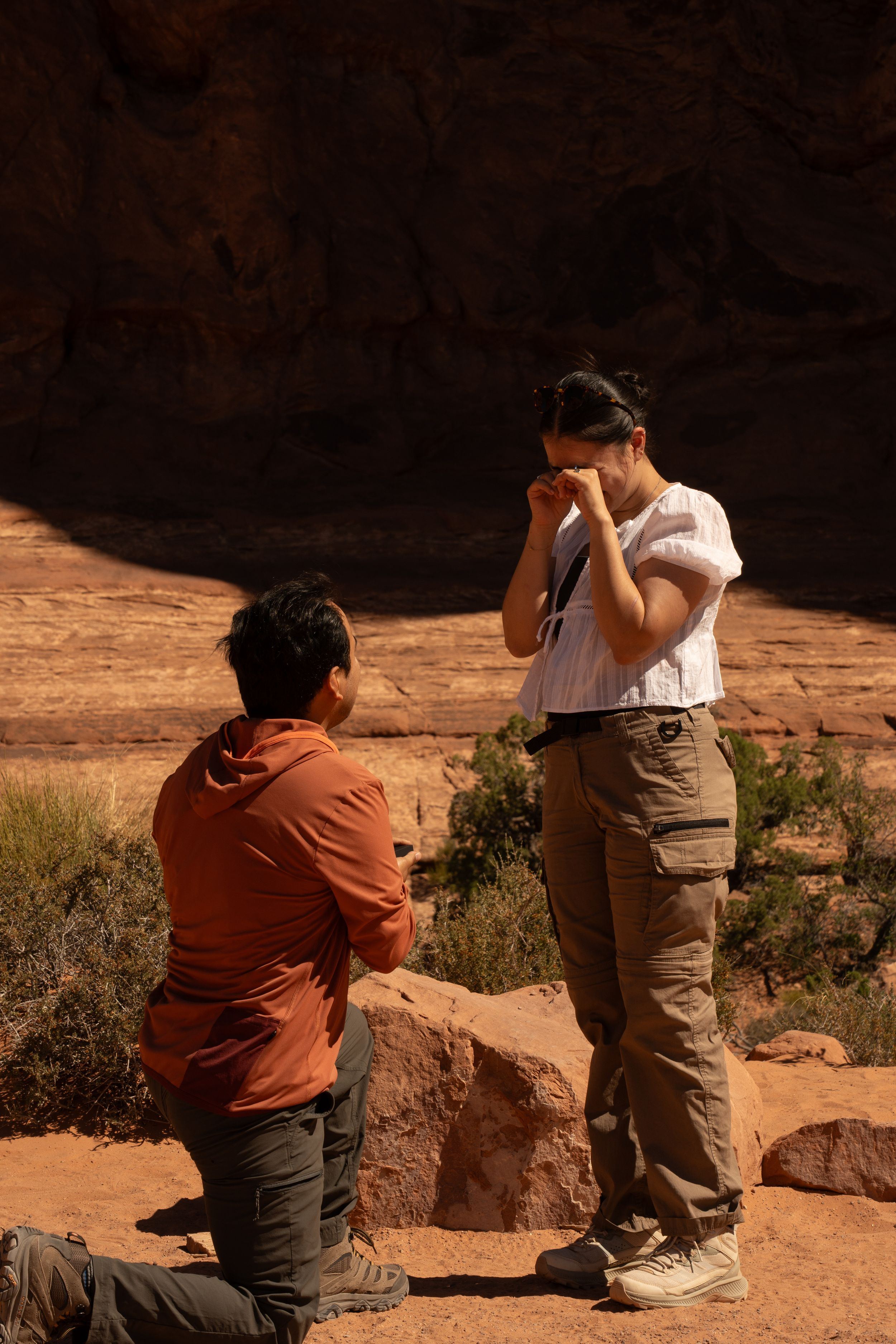 Man kneeling in proposal to emotional woman at red rock canyon landscape in sunny outdoor setting.