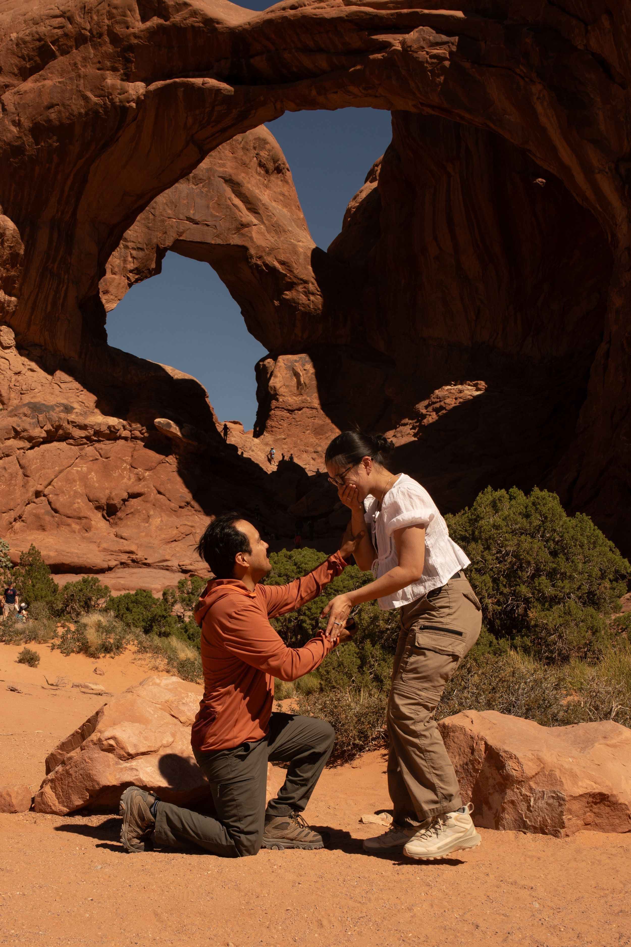Man proposing to woman on one knee at Double Arch in Arches National Park, Utah desert landscape.
