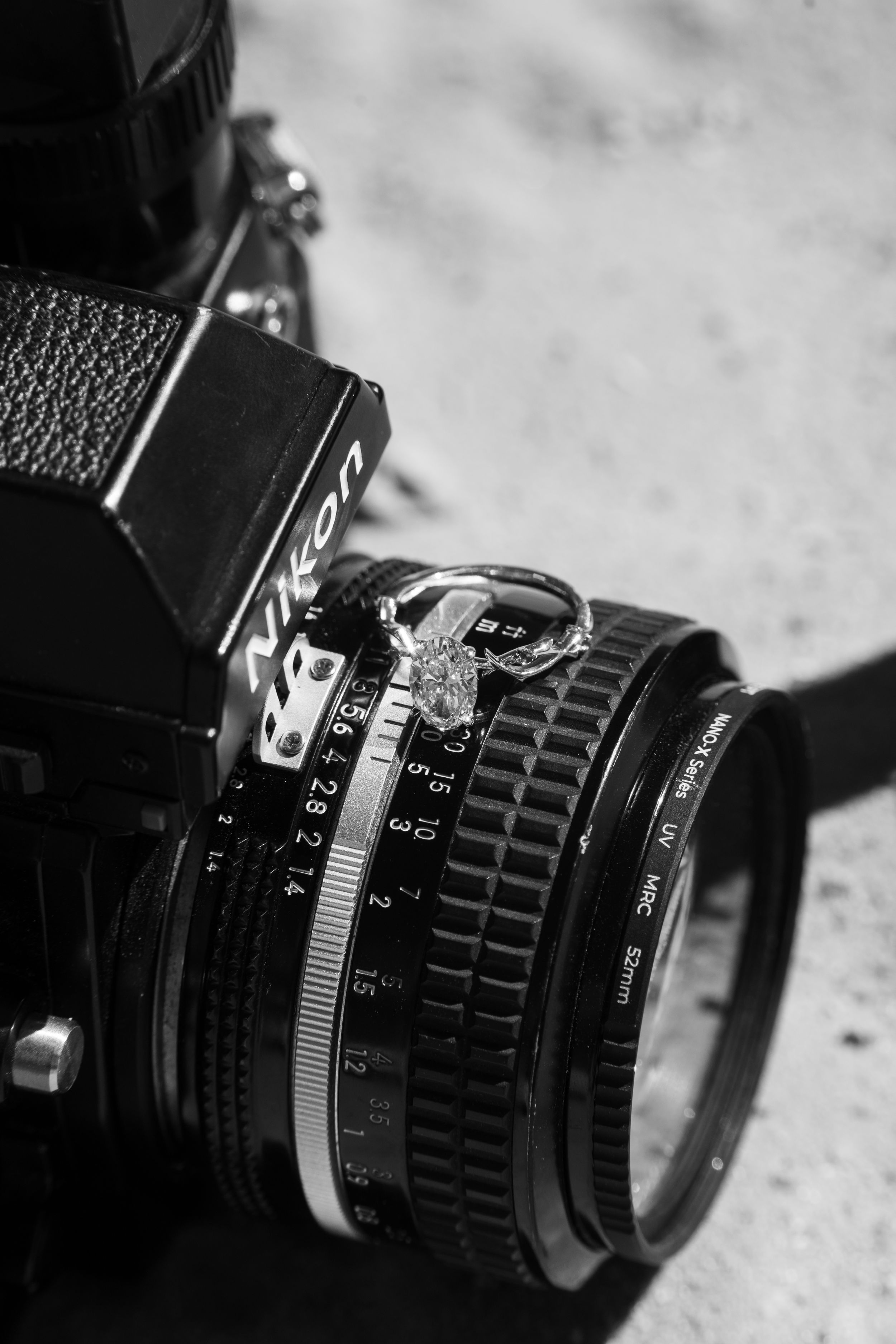 Black and white photo of a diamond engagement ring resting on a vintage Nikon camera lens.