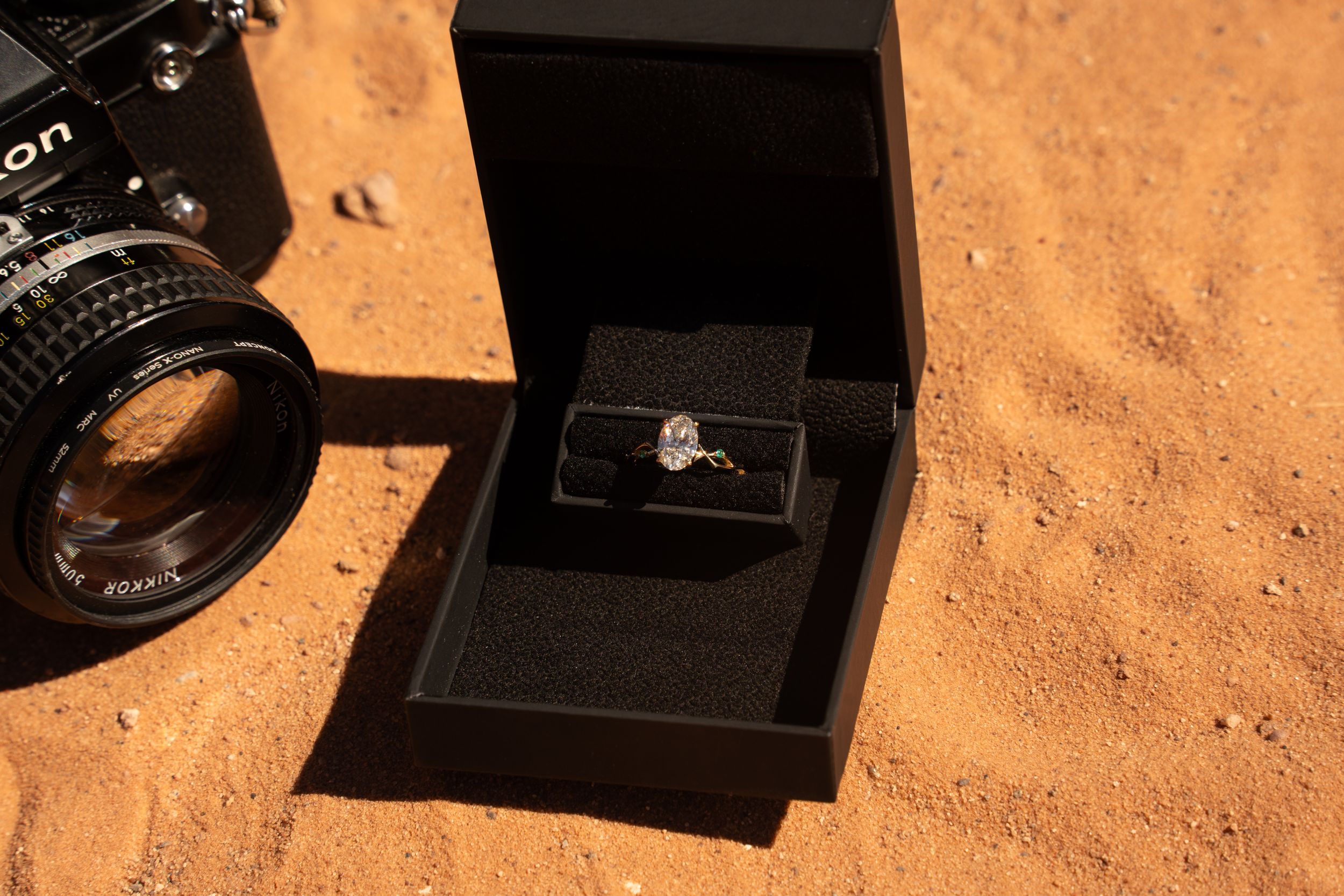 Diamond engagement ring in black box beside Nikon camera on red sand desert background.