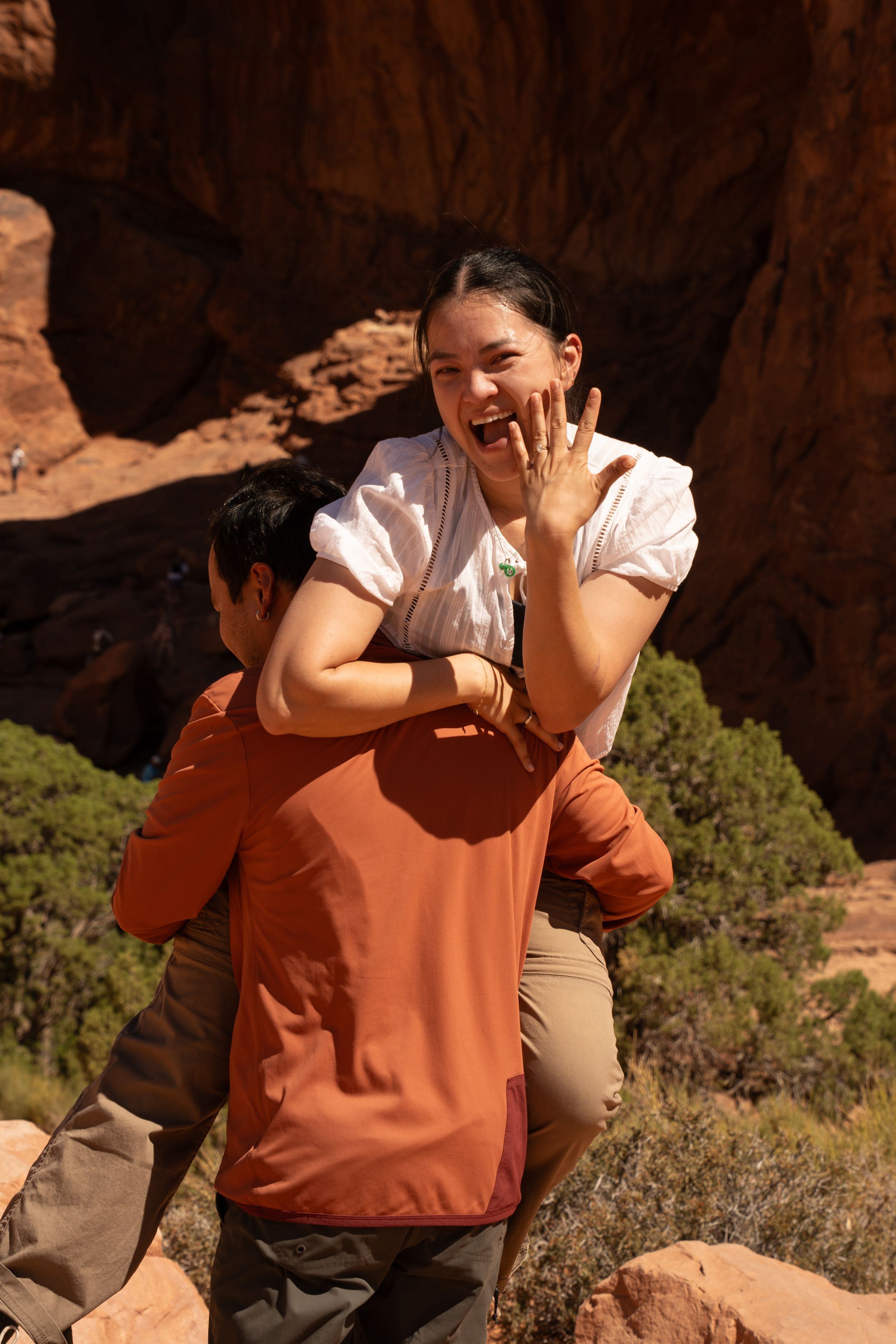 Woman showing engagement ring while being lifted by fiance amid red rock formations and arches.