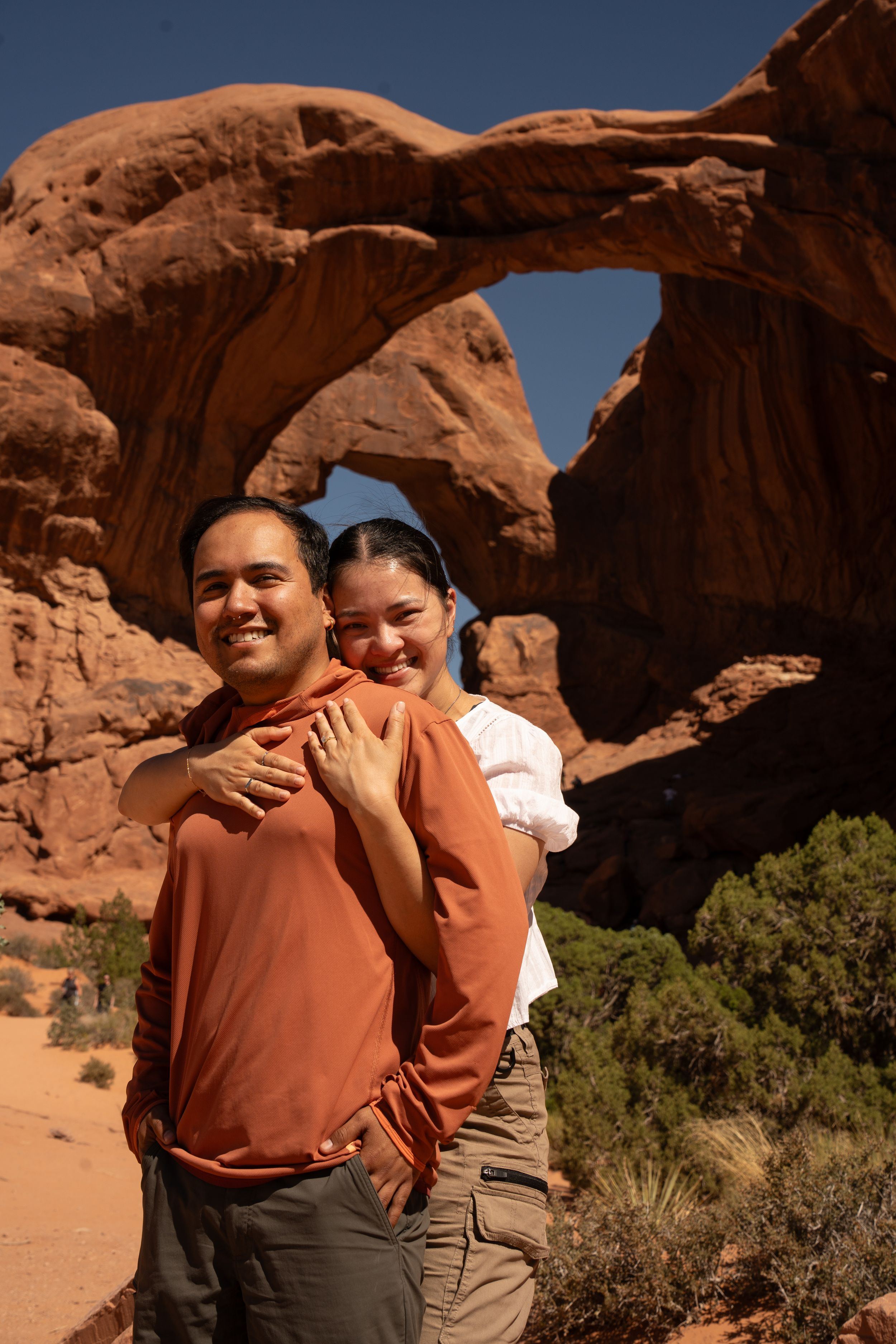 Smiling couple posing in front of a natural sandstone arch at Arches National Park on a sunny day and showing off engagement ring