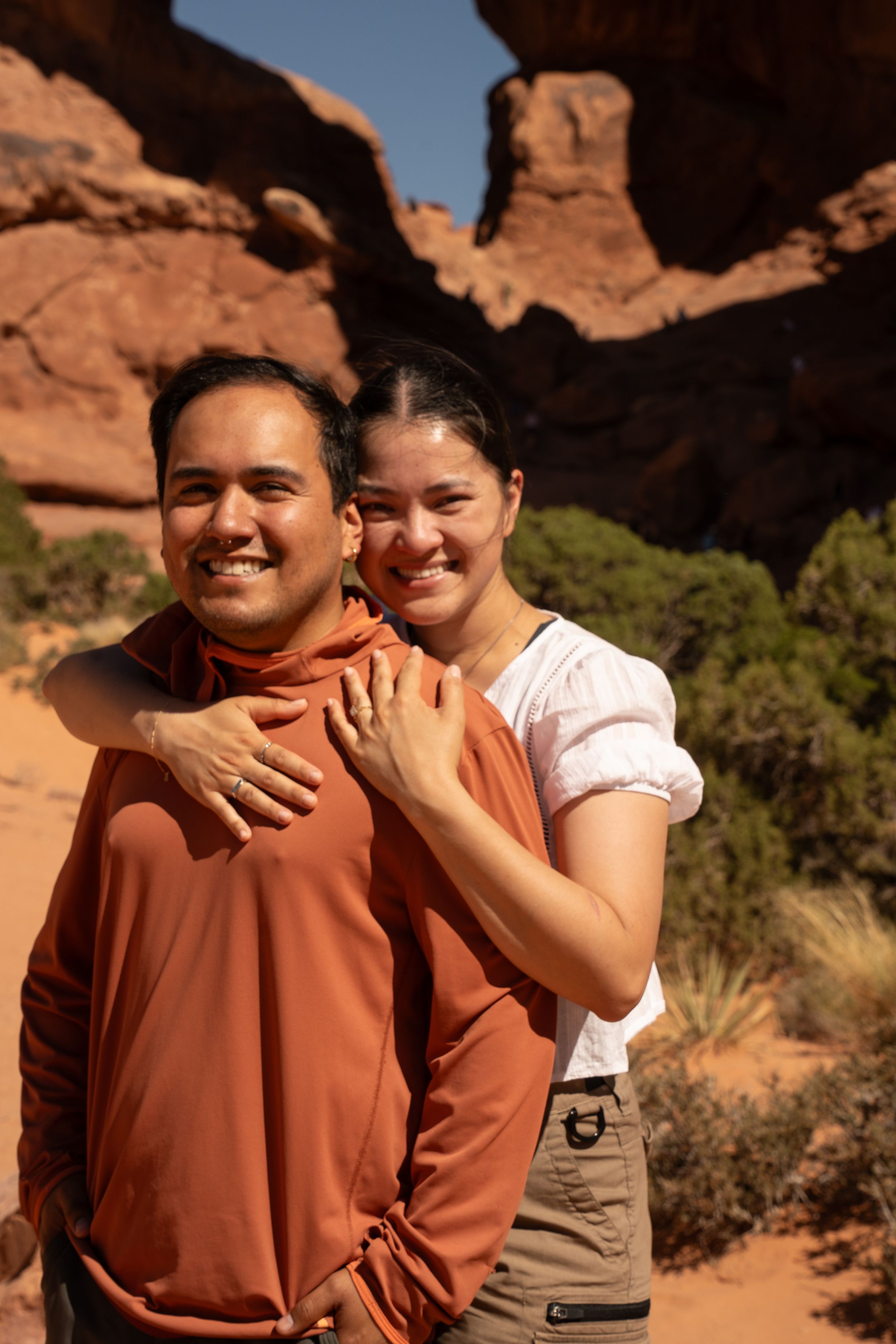 Smiling couple posing together showing off engagement ring in front of red rock formations in a desert landscape on a sunny day.