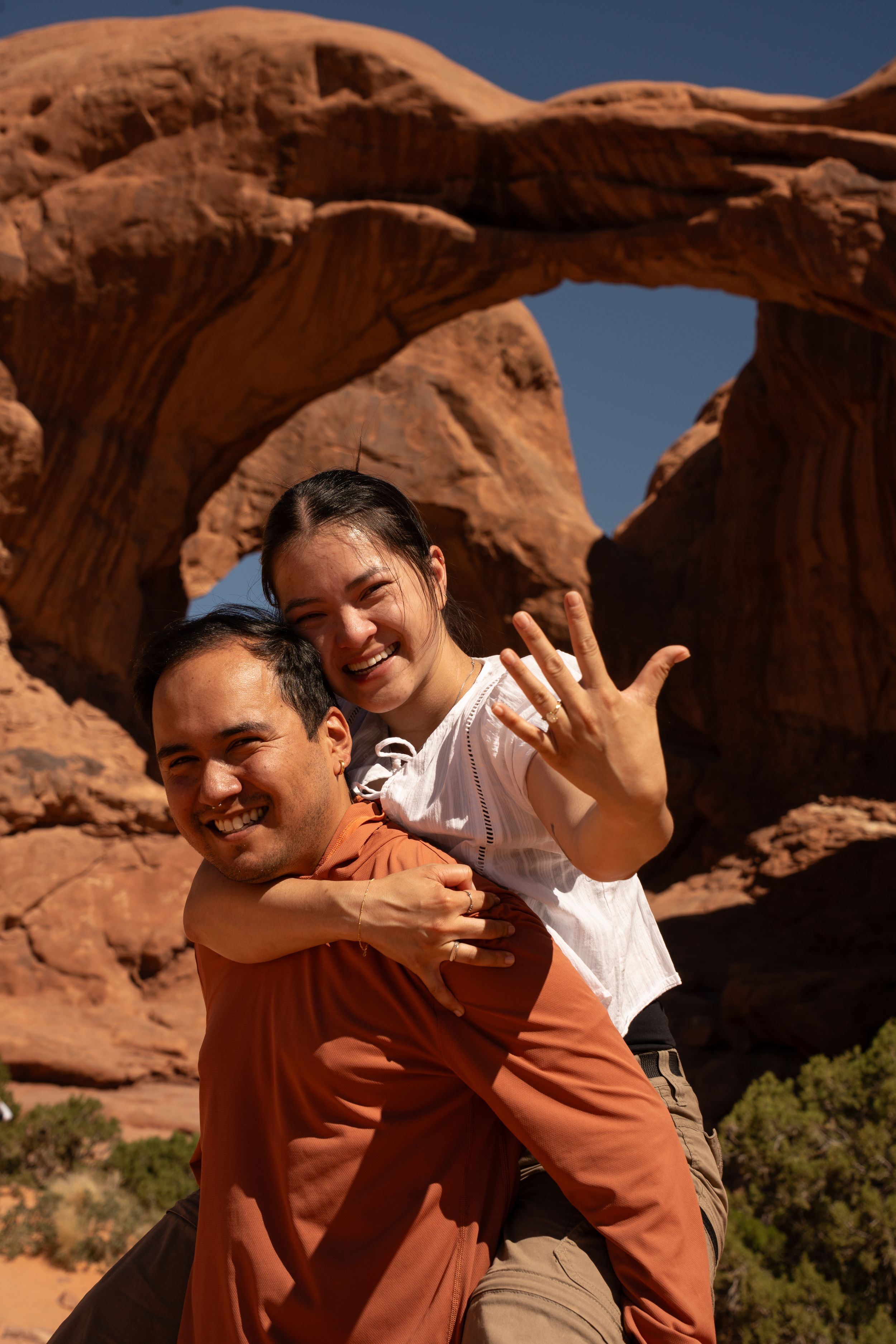 Couple celebrating engagement at Arches National Park, woman showing ring while piggyback riding man before red rock arch.