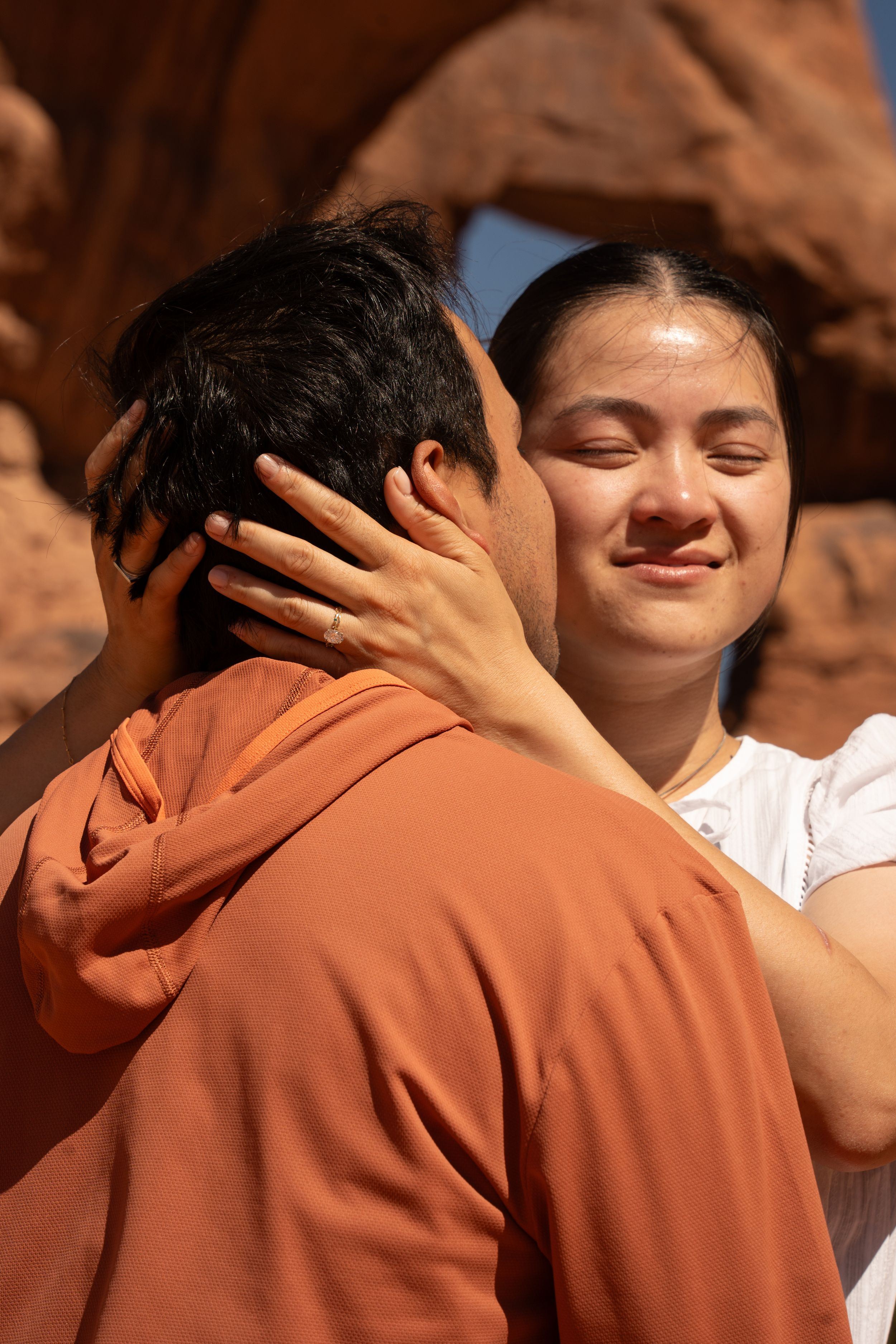 A smiling Asian woman with closed eyes tenderly holds a man's head while embracing outdoors near red rock formations.