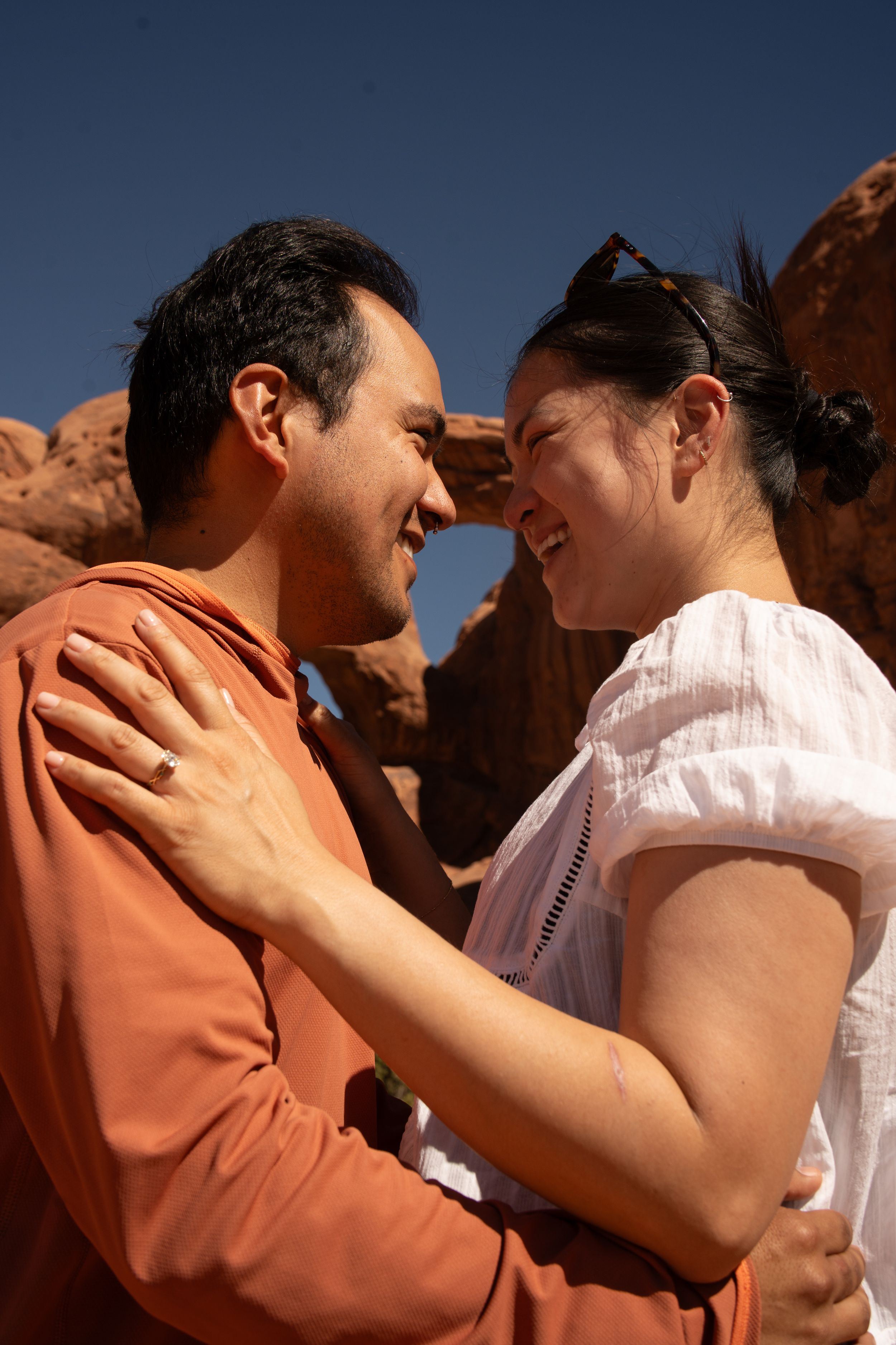 Happy couple embracing and smiling at each other in front of red rock formations under a clear blue sky.