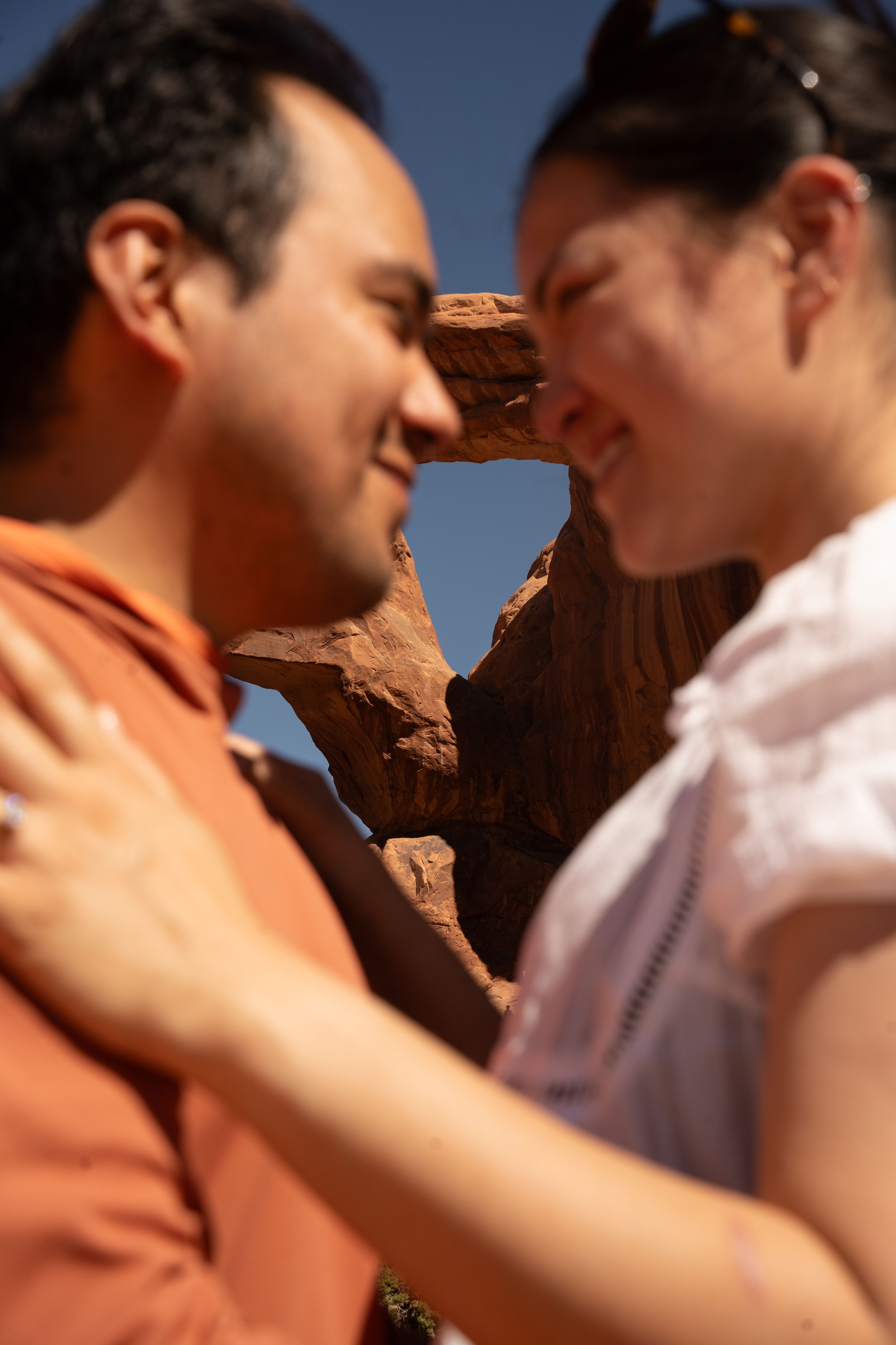 engaged Couple smiling at each other with a red rock arch formation in the background under a clear blue sky.