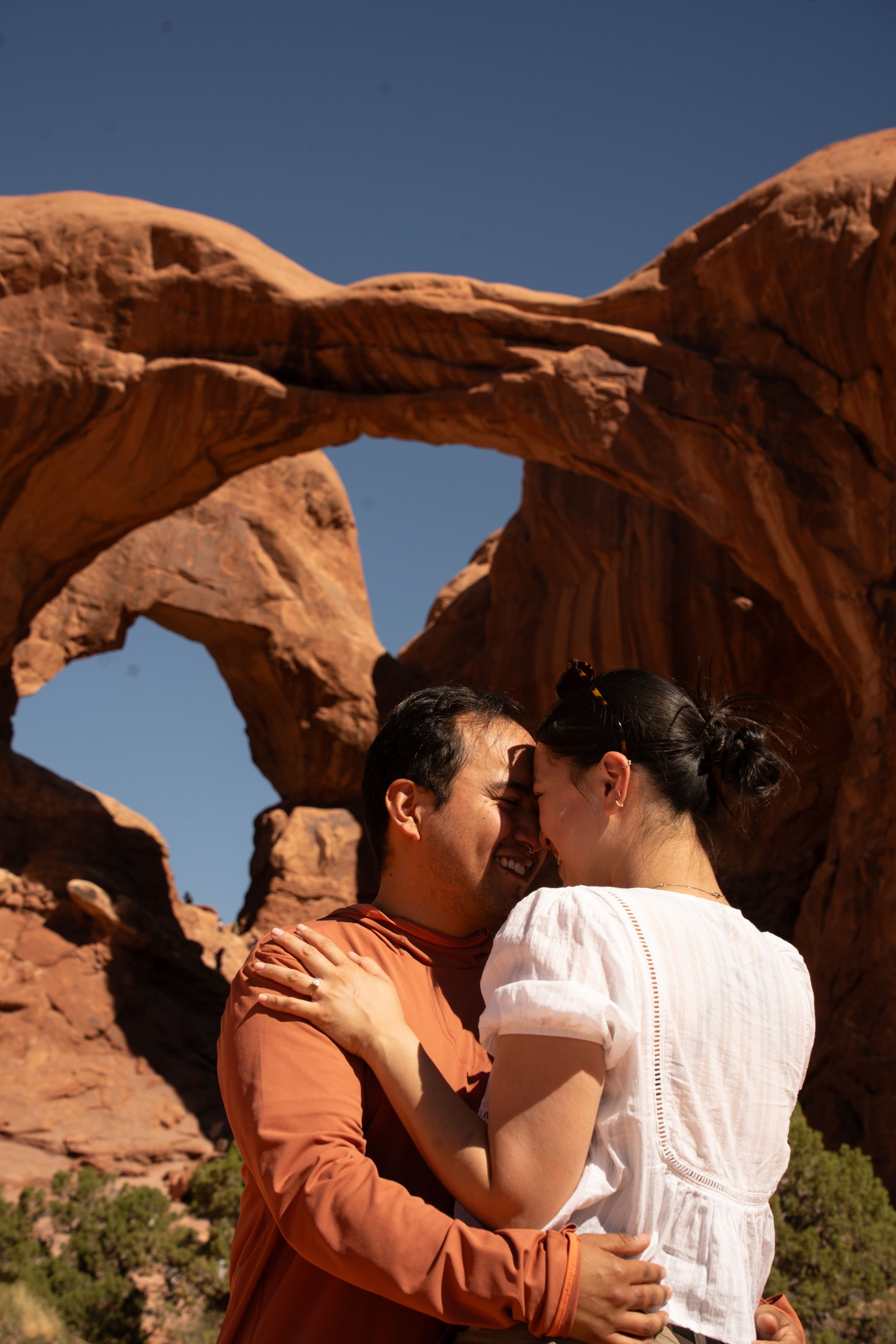 Couple embracing and kissing in front of Double Arch at Arches National Park, Utah, with red sandstone formations.