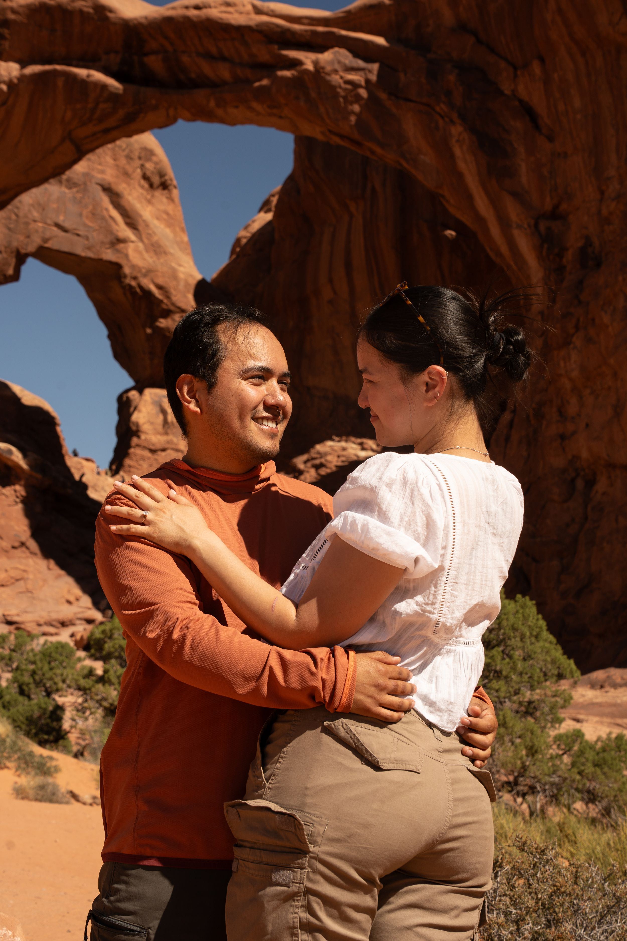 Couple embracing and smiling before a natural sandstone arch at Arches National Park, Utah.