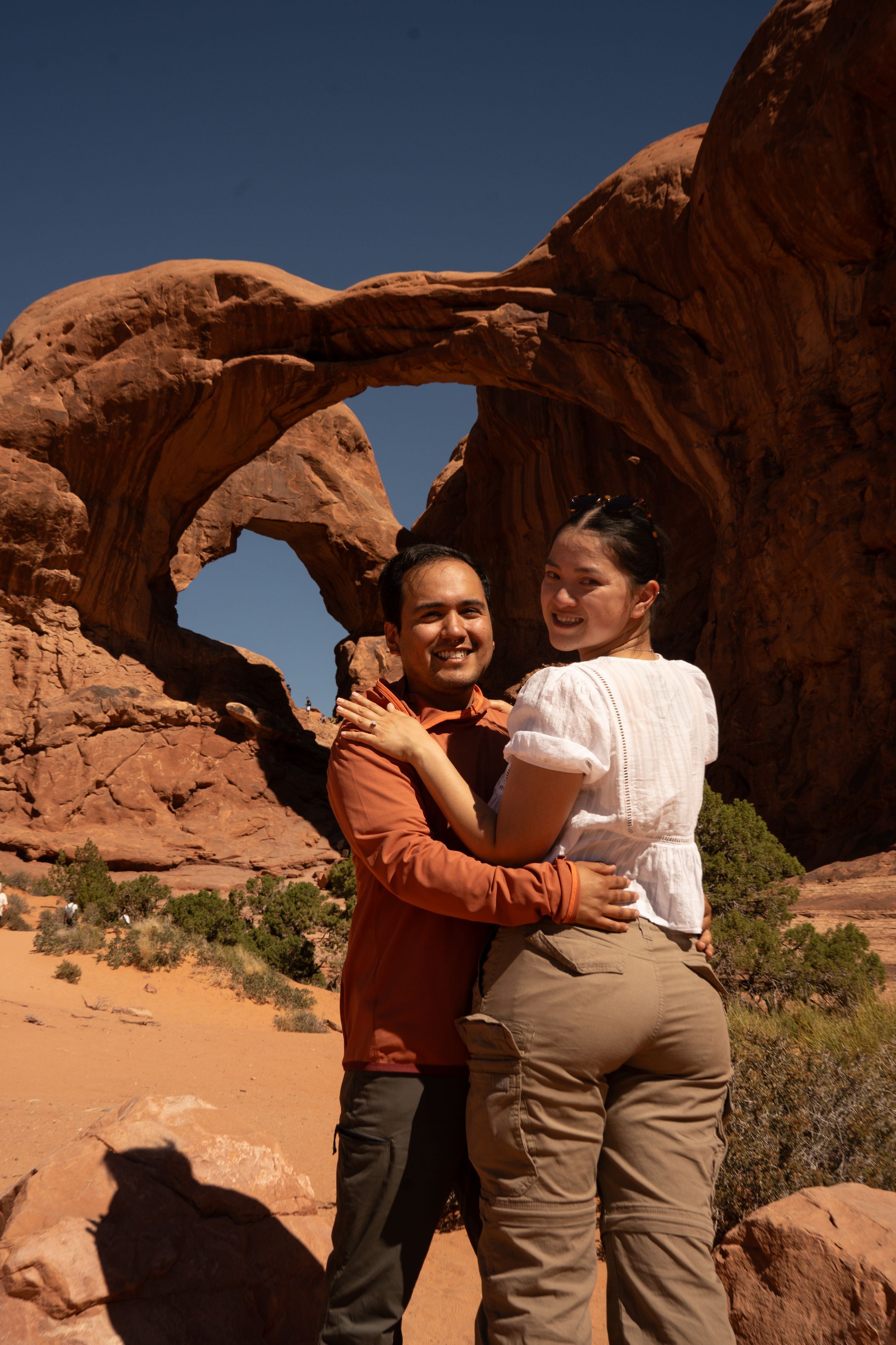 Couple embracing and smiling in front of a natural sandstone arch at Arches National Park on a sunny day.