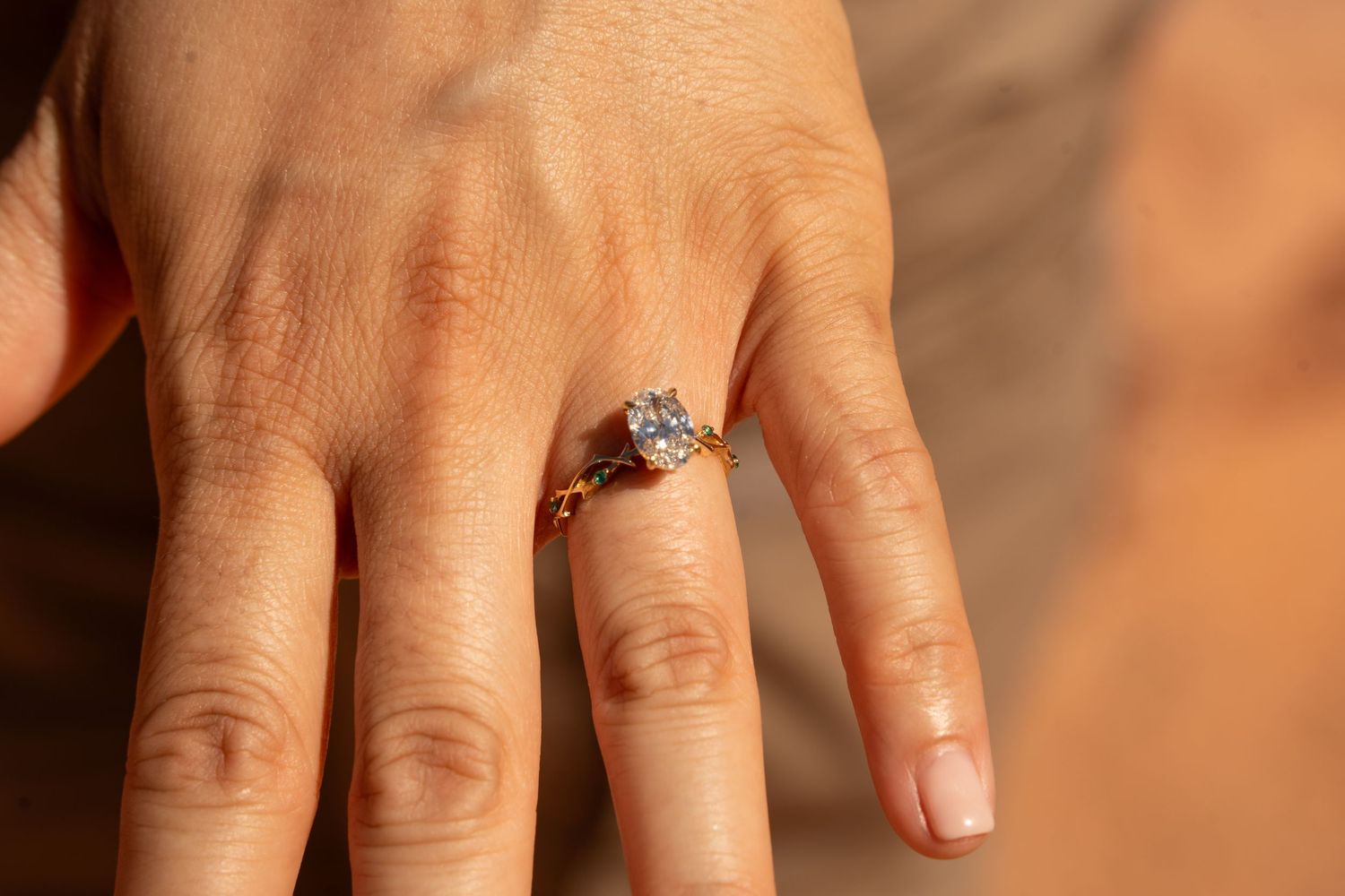 Close-up of a hand wearing a gold engagement ring with a large oval diamond solitaire and small green accent stones.